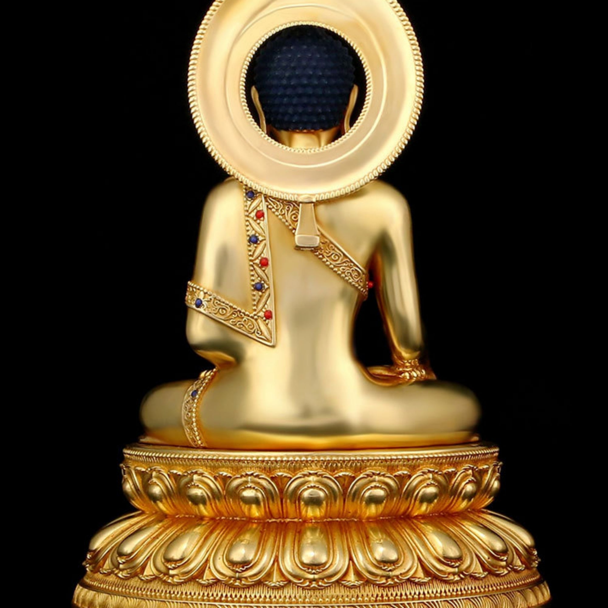 A golden Buddha statue shown from the back, seated on an ornate lotus-style base with a large circular halo behind the head and small multicolor jewel accents on the garment, against a solid black background.