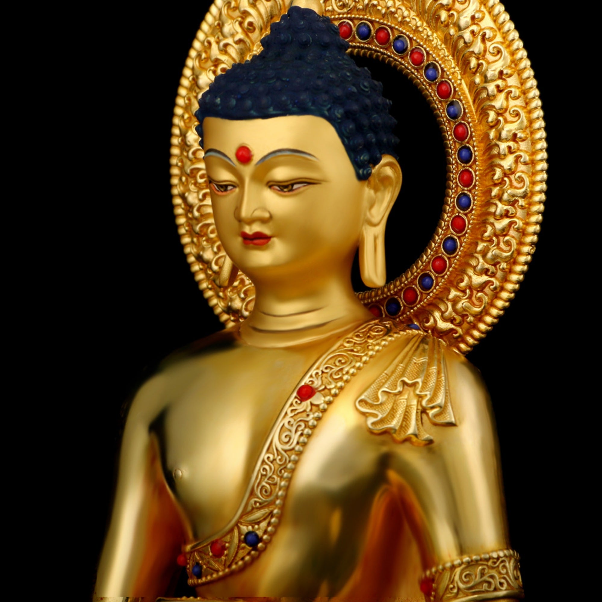 A close-up studio shot of a gold-toned Buddha statue with a blue hair and small red facial ornaments, set against a solid black background.