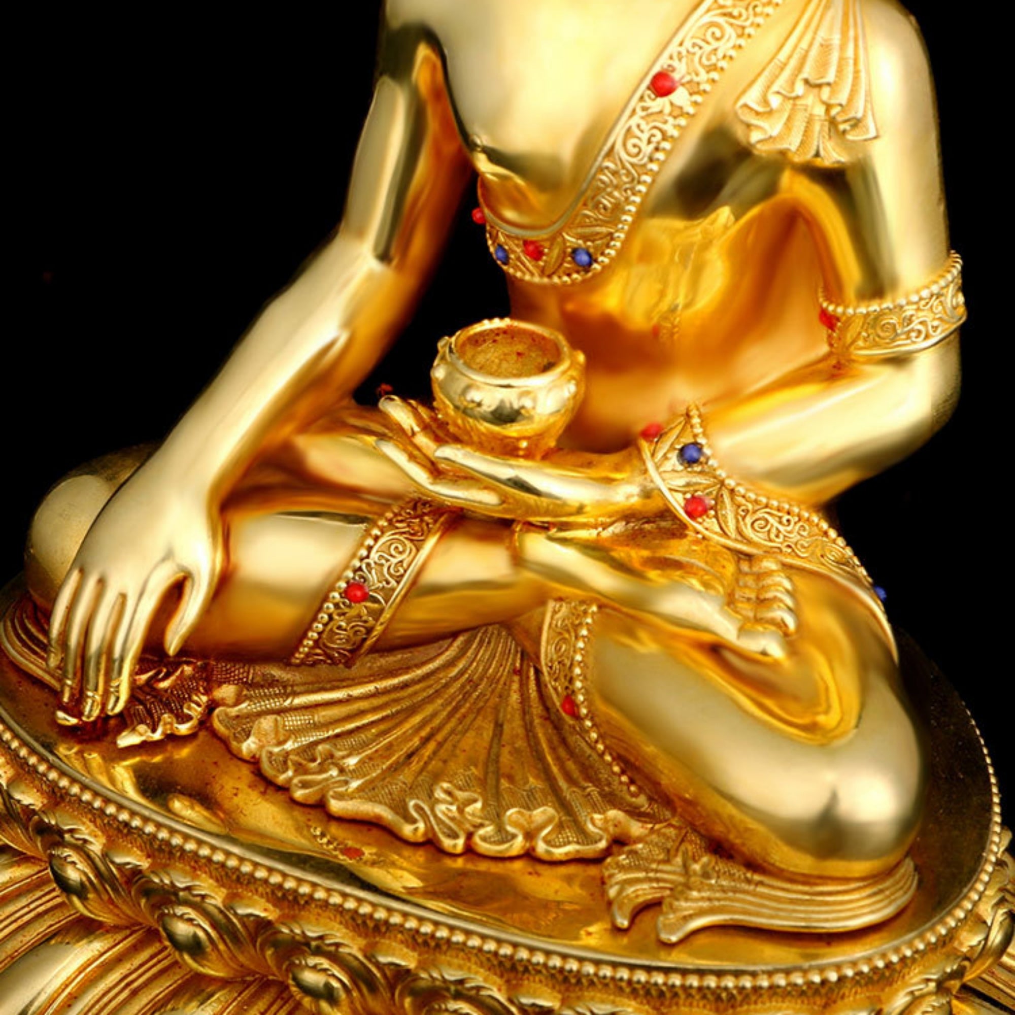 A close-up of a polished gold-toned figurine depicting a seated human figure with ornate jewelry and red gem accents against a black background.
