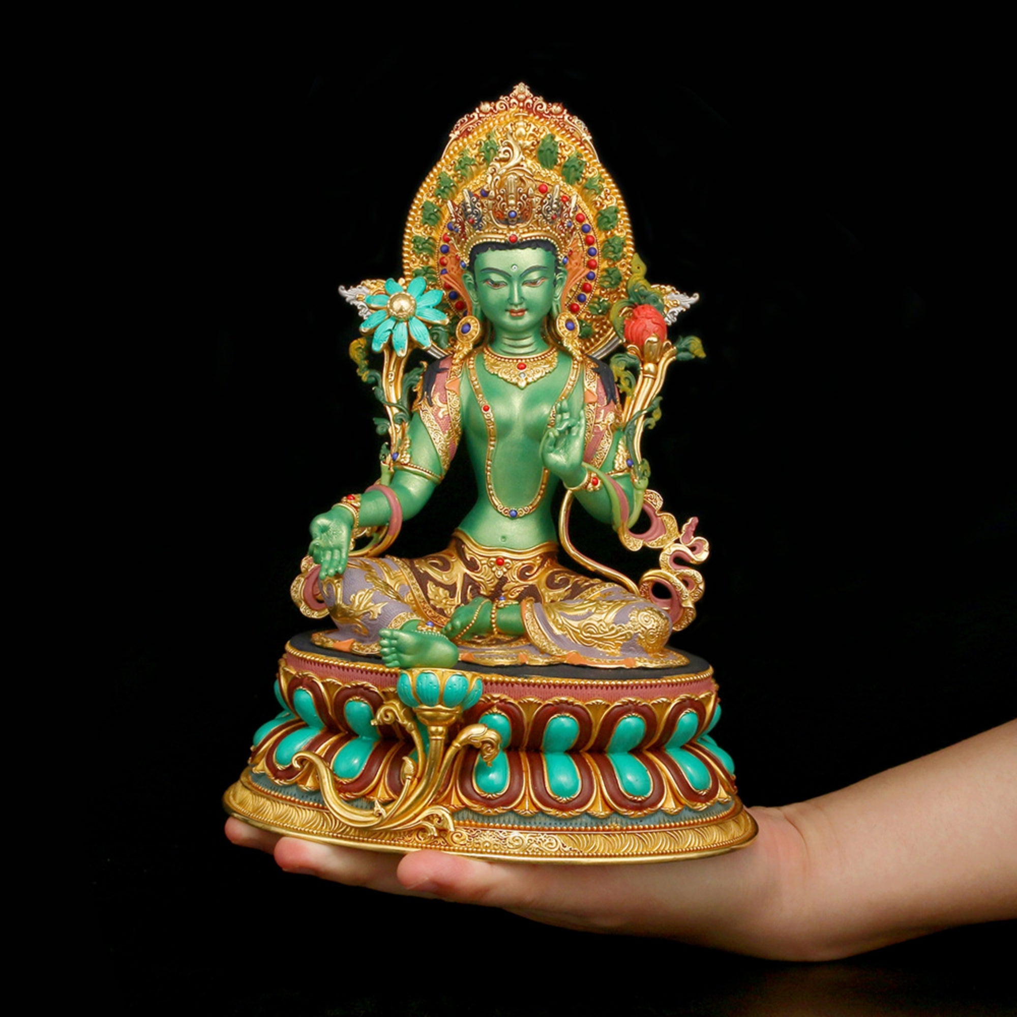 A green seated Buddha statue with ornate gold, turquoise, and red detailing, displayed on a person's hand against a solid black background.