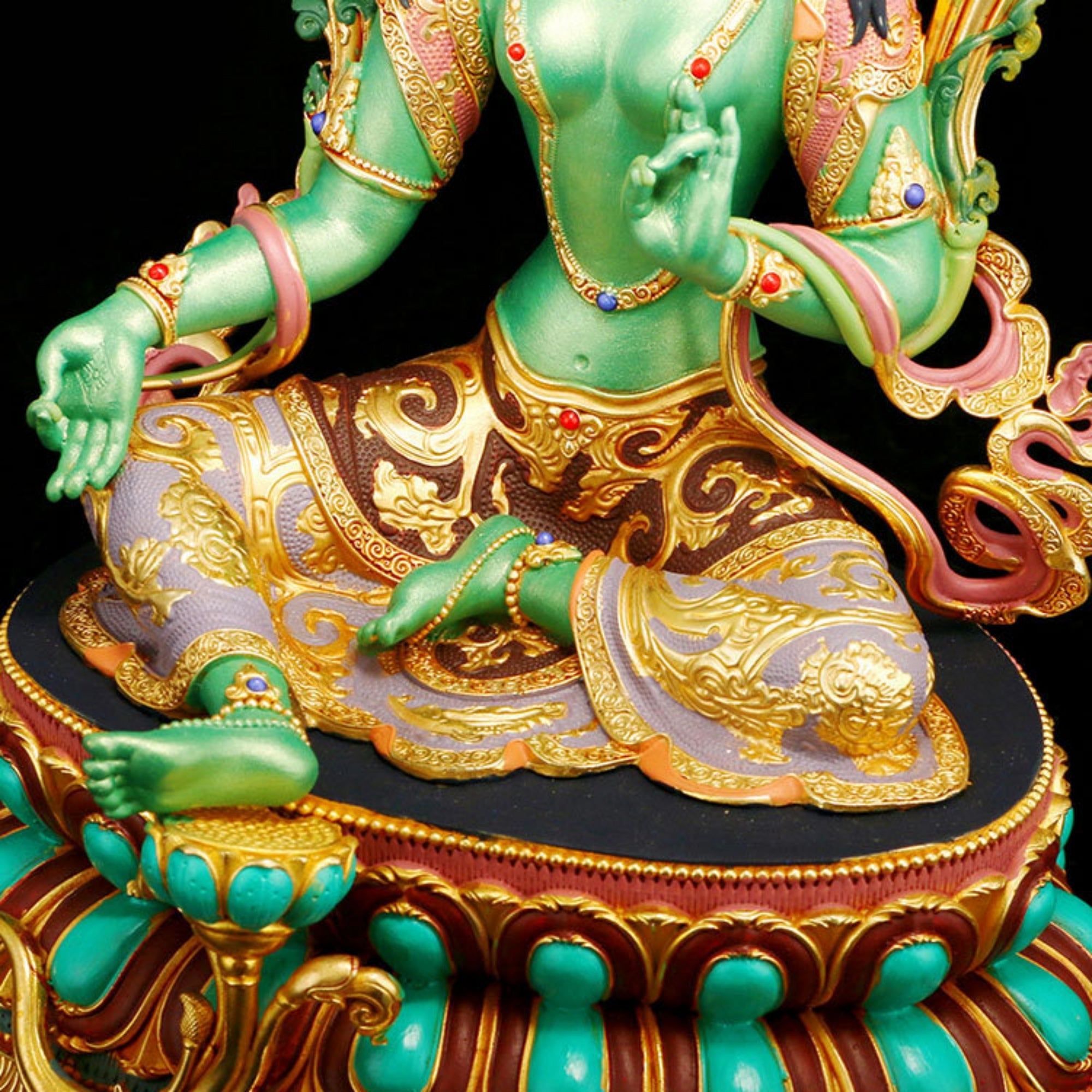 A close-up of a seated green deity statue with ornate gold detailing, pink drapery, turquoise accents, and a black base against a black background.