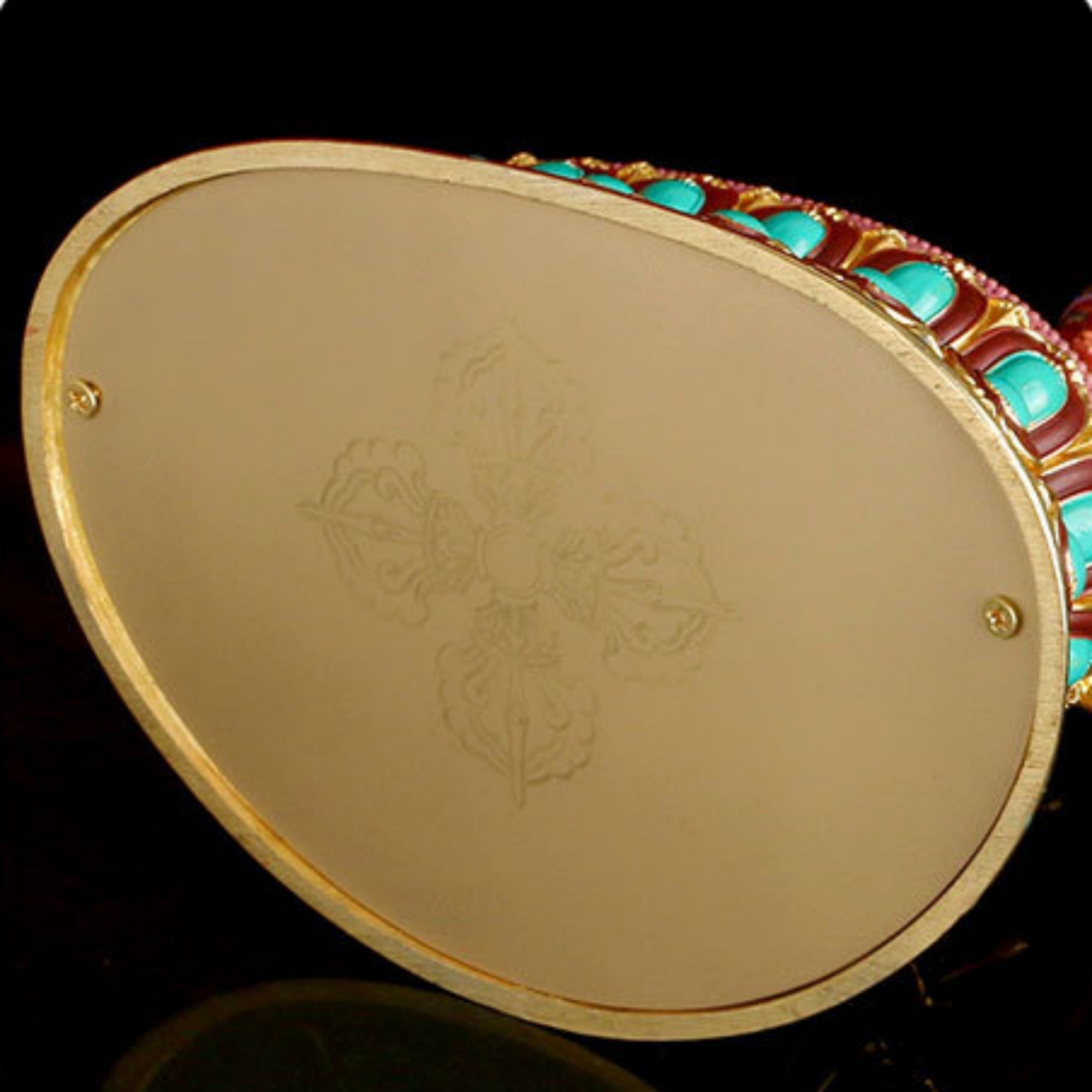 An oval gold-toned pendant or medallion with a faint engraved cross design on the front, bordered by turquoise and red gemstone accents on a black background.