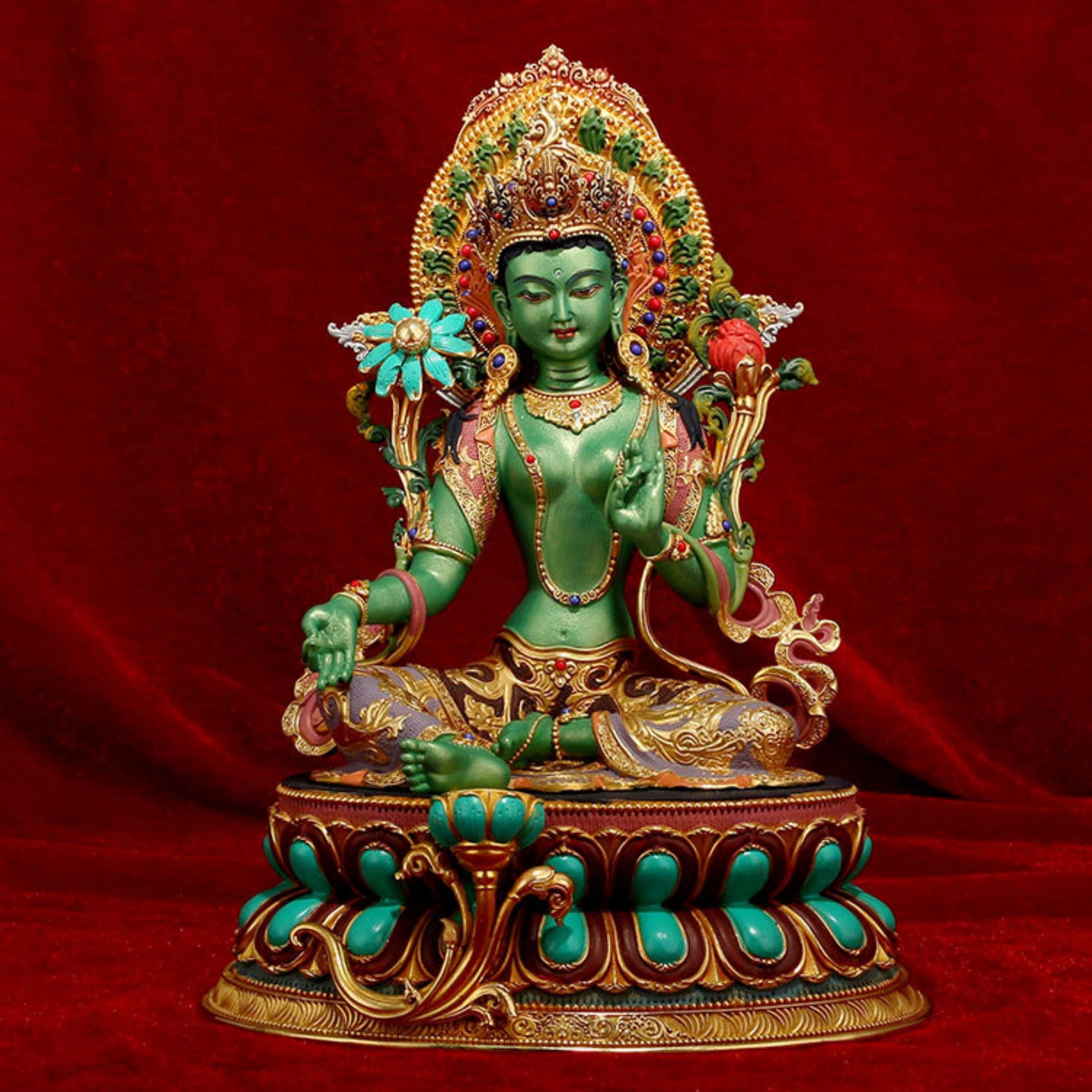 A green seated deity statue with ornate gold detailing, turquoise accents, and a red floral crown, posed on a lotus base against a red fabric backdrop.