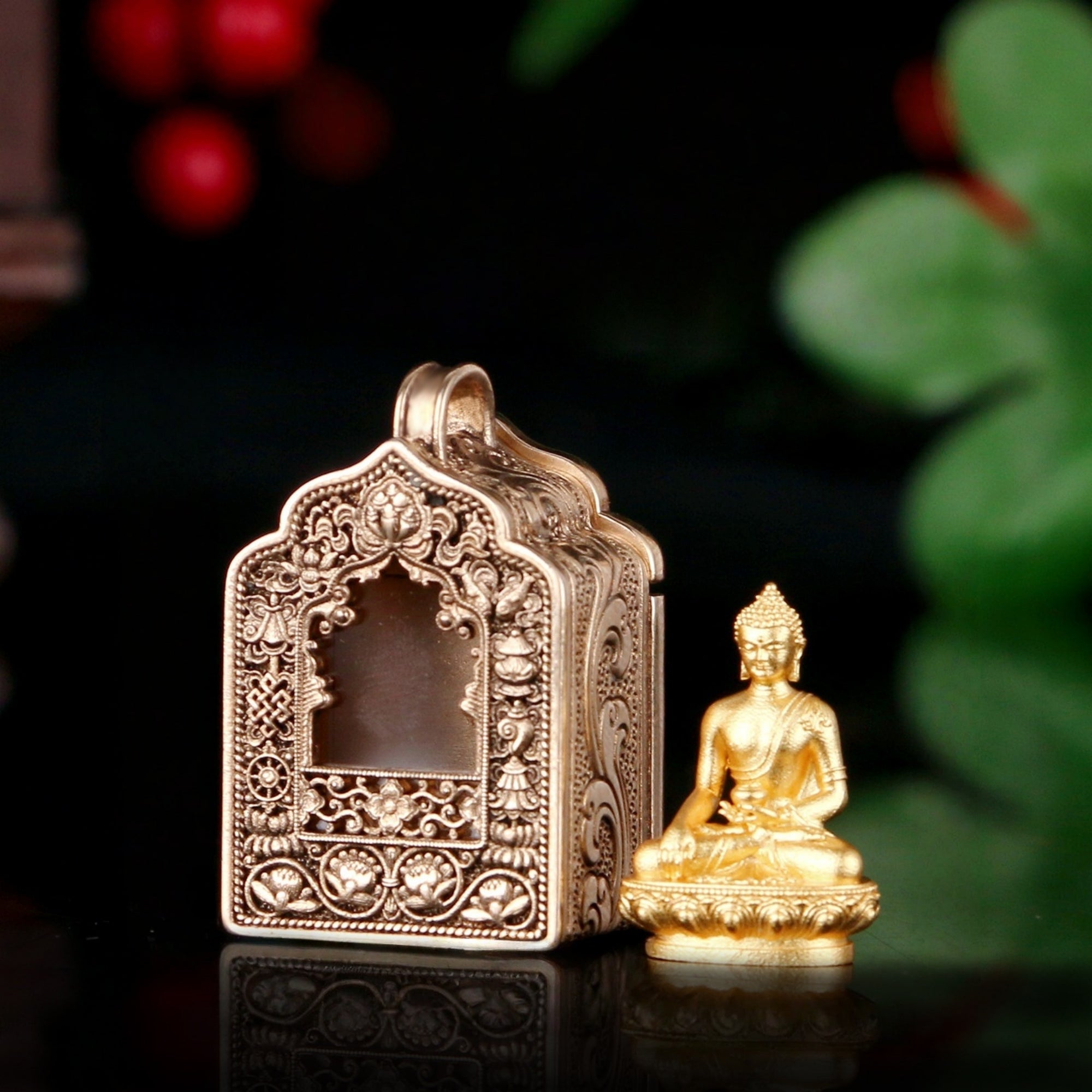 An ornate gold-toned metal box with intricate filigree detailing and a small seated Buddha figurine beside it on a dark reflective surface, with blurred green leaves and red berries in the background.