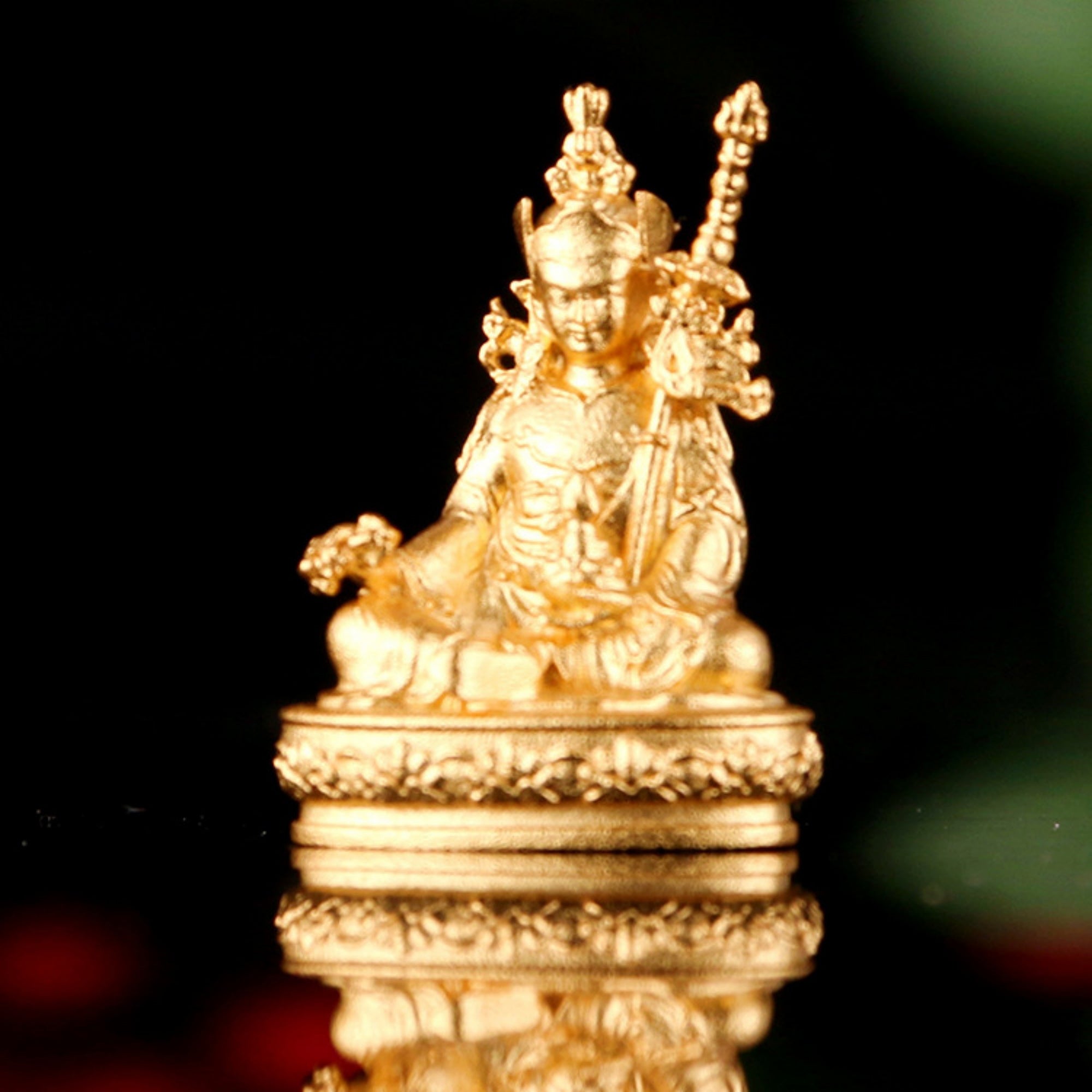 A small golden seated deity statue on an ornate circular base, photographed against a dark black background with a soft reflection below.