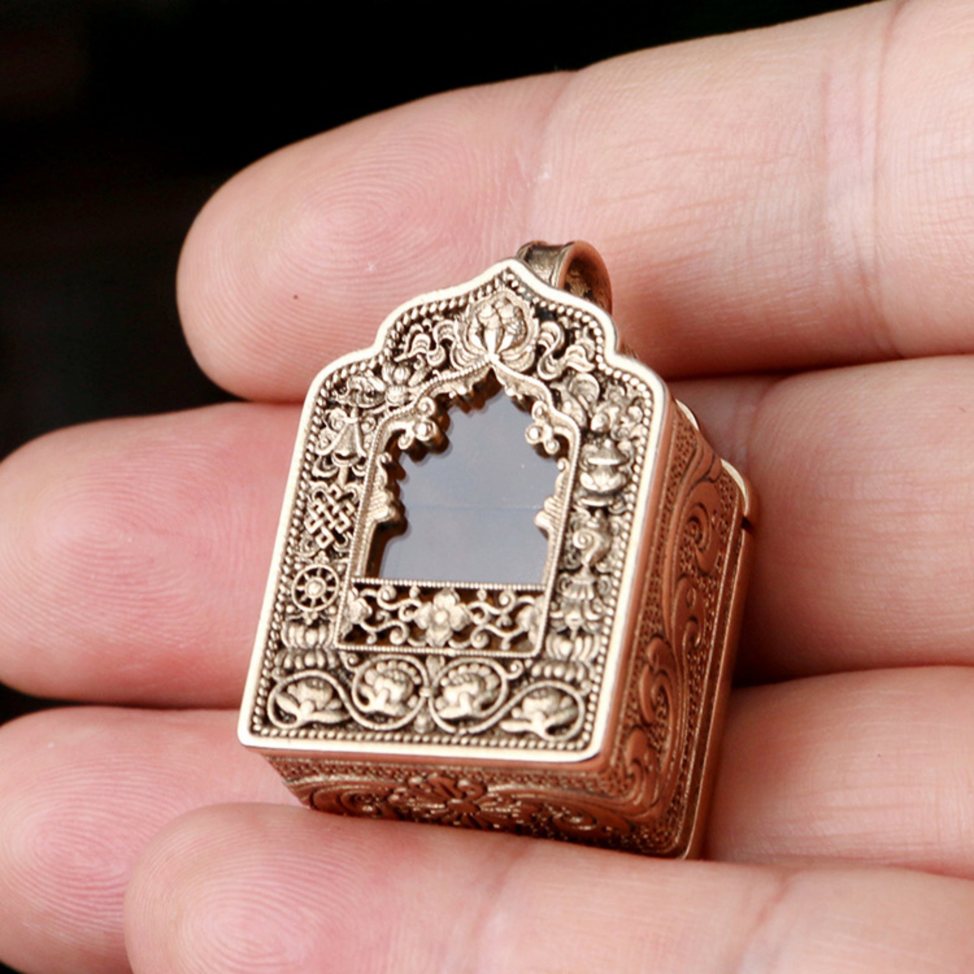 An ornate gold-toned pendant with intricate filigree detailing and a central arch cutout, held in a person's fingers against a dark background.