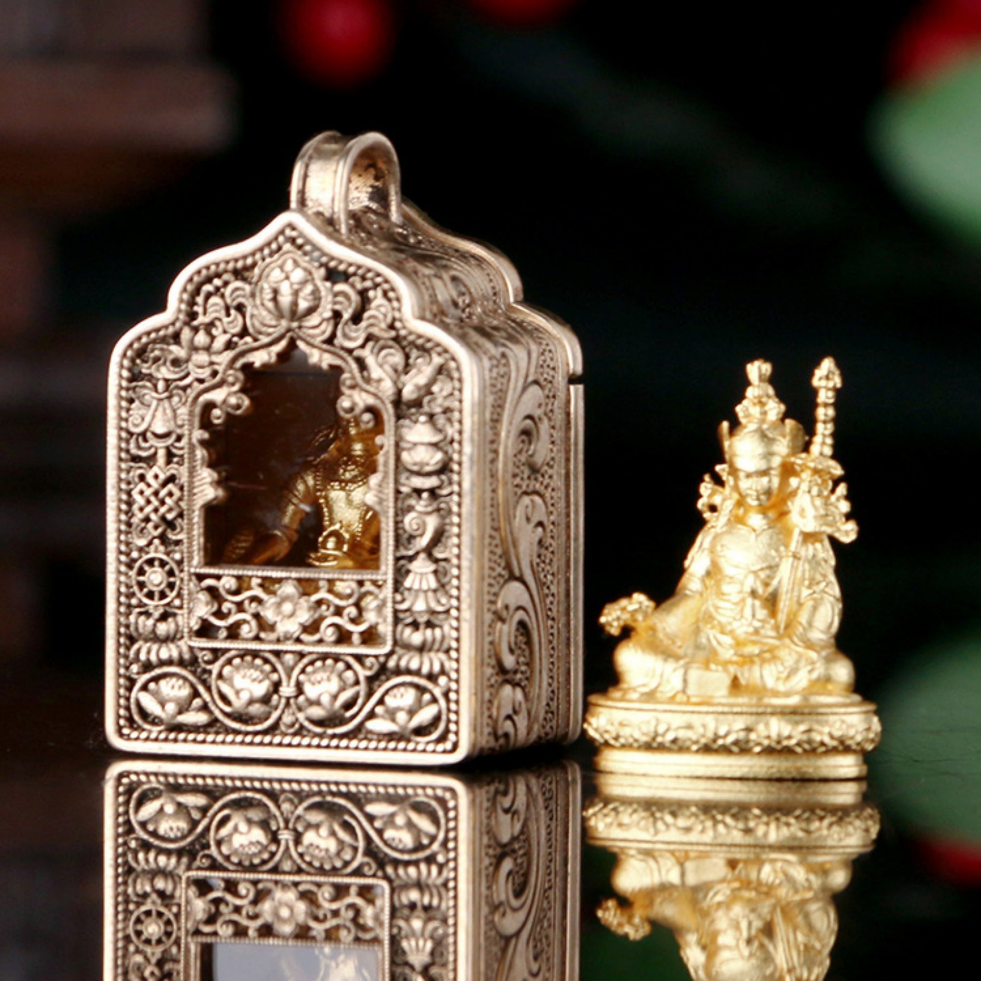 An ornate silver-toned metal miniature temple box with intricate filigree detailing and a small window, shown beside a gold-toned seated deity figurine on a reflective dark surface.