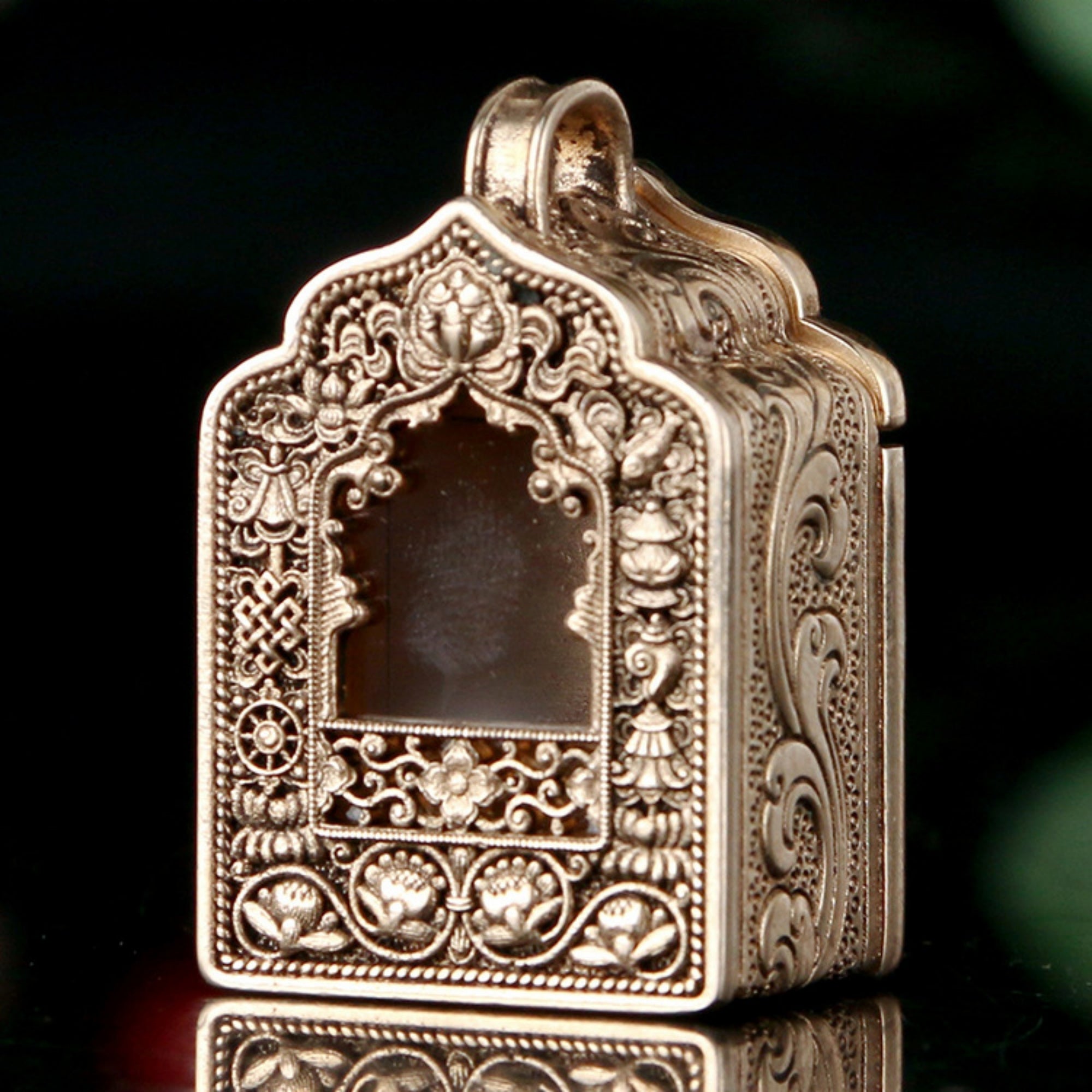 An ornate metallic pendant or locket with intricate floral and scroll engravings, shown on a dark reflective surface against a dark background.