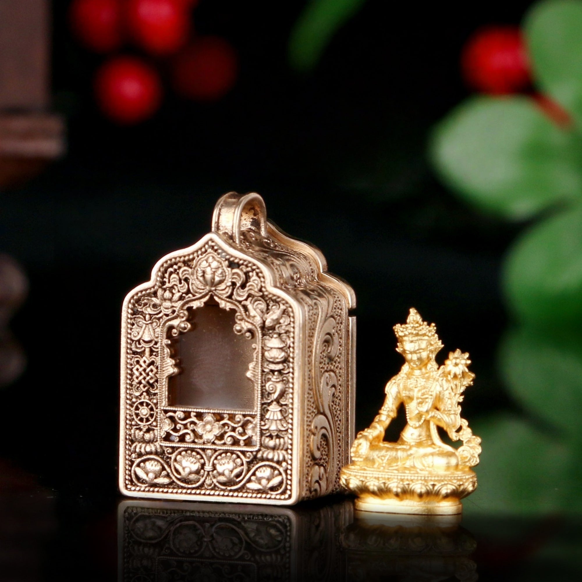 An ornate gold-toned metal miniature temple with intricate relief carvings, shown beside a small seated gold figurine on a dark reflective surface with blurred red berries and green leaves in the background.