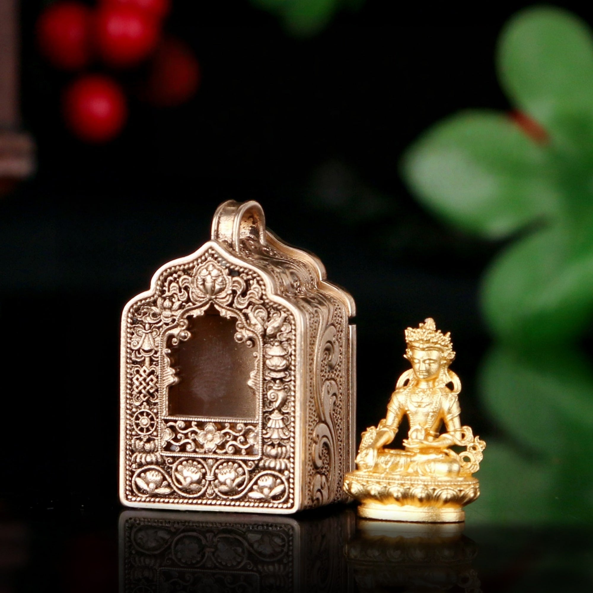 An ornate antique-style gold-toned metal box with intricate filigree detailing and a small seated deity figurine beside it, set against a dark background with blurred red berries and green leaves.