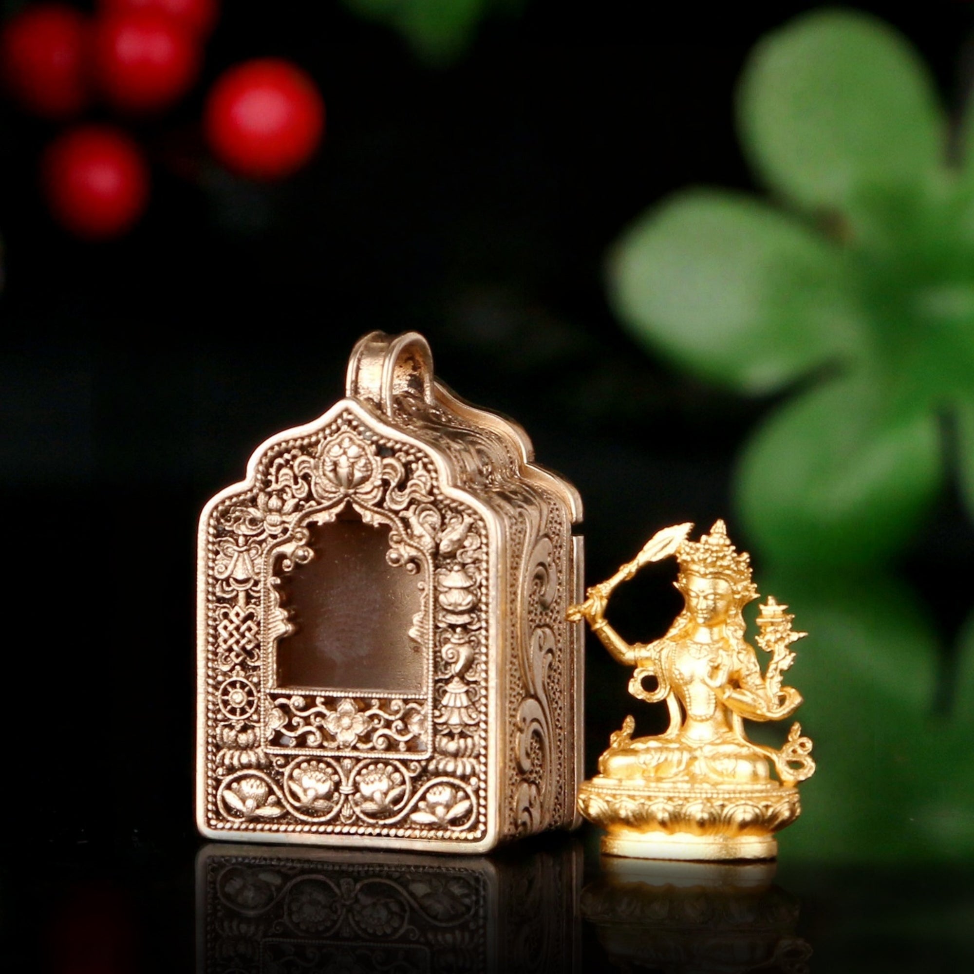 An ornate gold-toned metal box with intricate relief carvings, shown next to a small golden deity figurine on a dark reflective surface with blurred green leaves and red berries in the background.