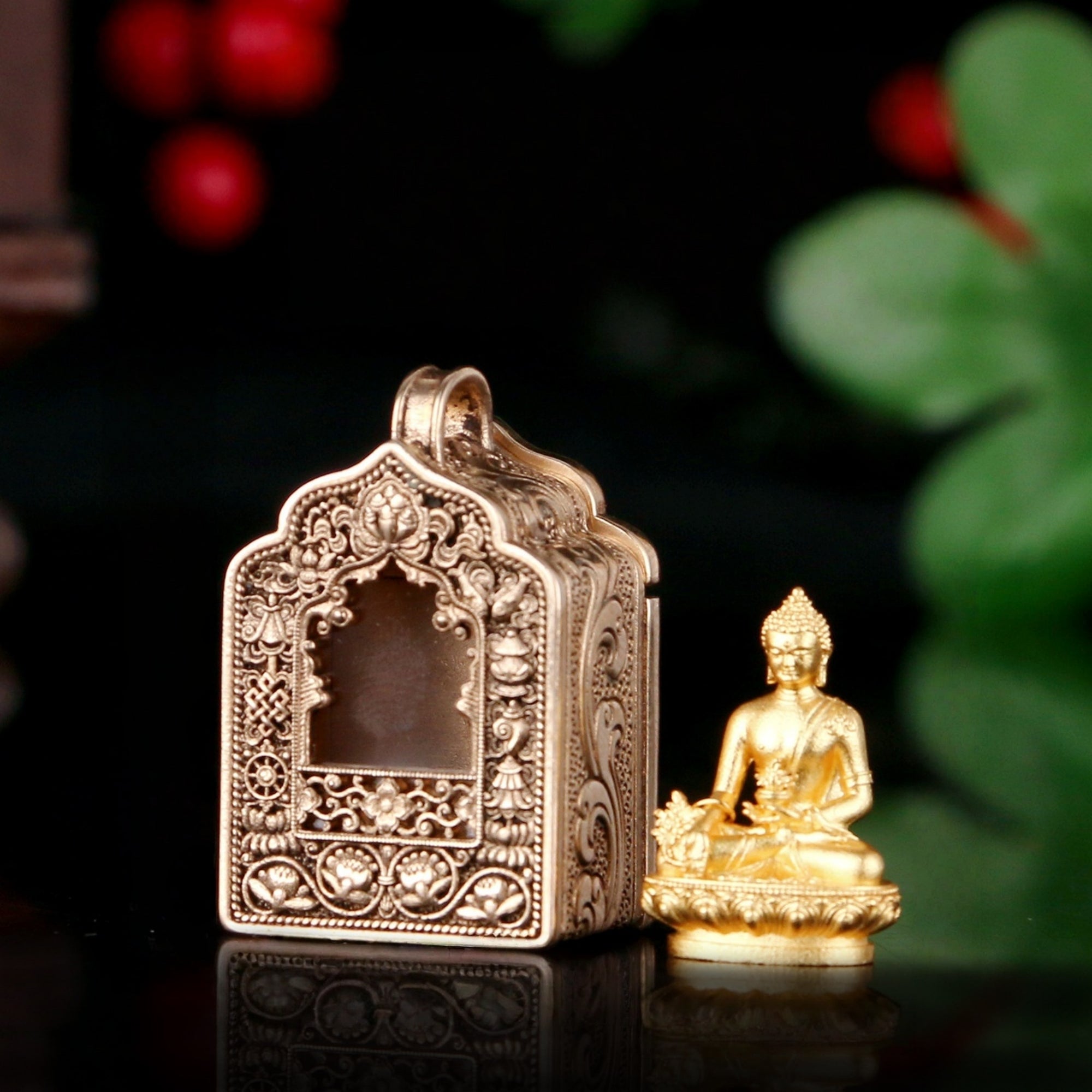 An ornate metallic temple-shaped pendant or box with intricate filigree detailing, shown next to a small gold Buddha statue on a dark reflective surface with blurred green leaves and red berries in the background.