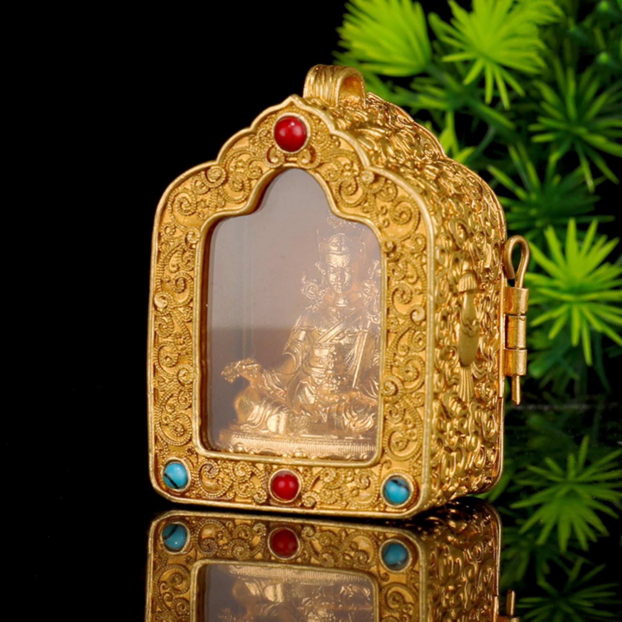 An ornate gold-toned decorative box or pendant with carved scrollwork, a transparent front panel showing a small figurine inside, and small red and blue gemstone accents, photographed against a black background with green foliage at the side.