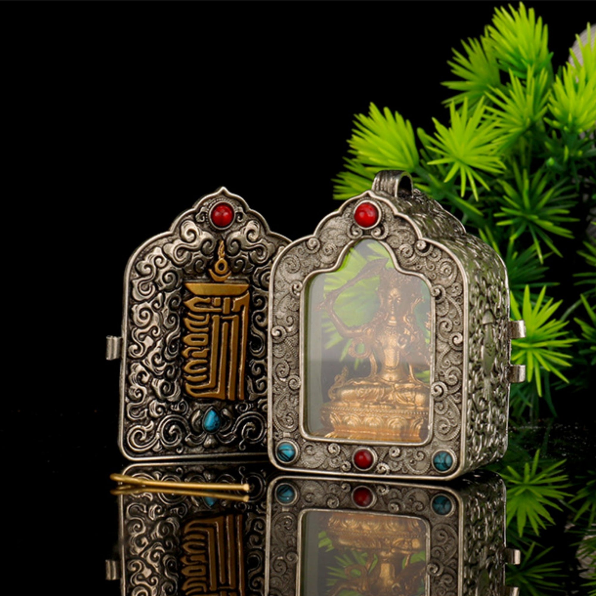 Two ornate silver-toned metal boxes with intricate scrollwork and red and blue gemstone accents, displayed on a reflective black surface with green foliage in the background.