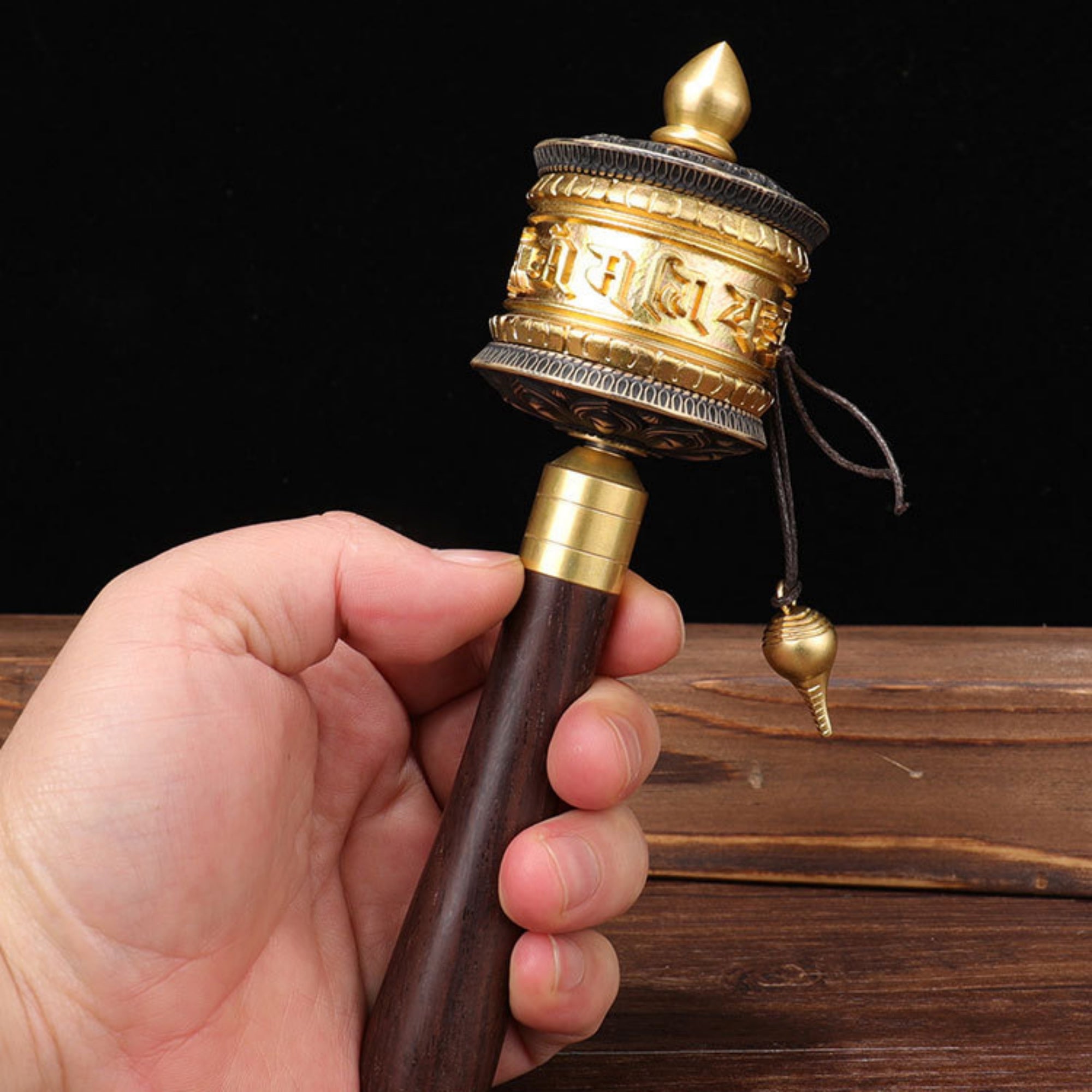 Hand holding a traditional prayer wheel with gold and brown colors on a wooden surface.