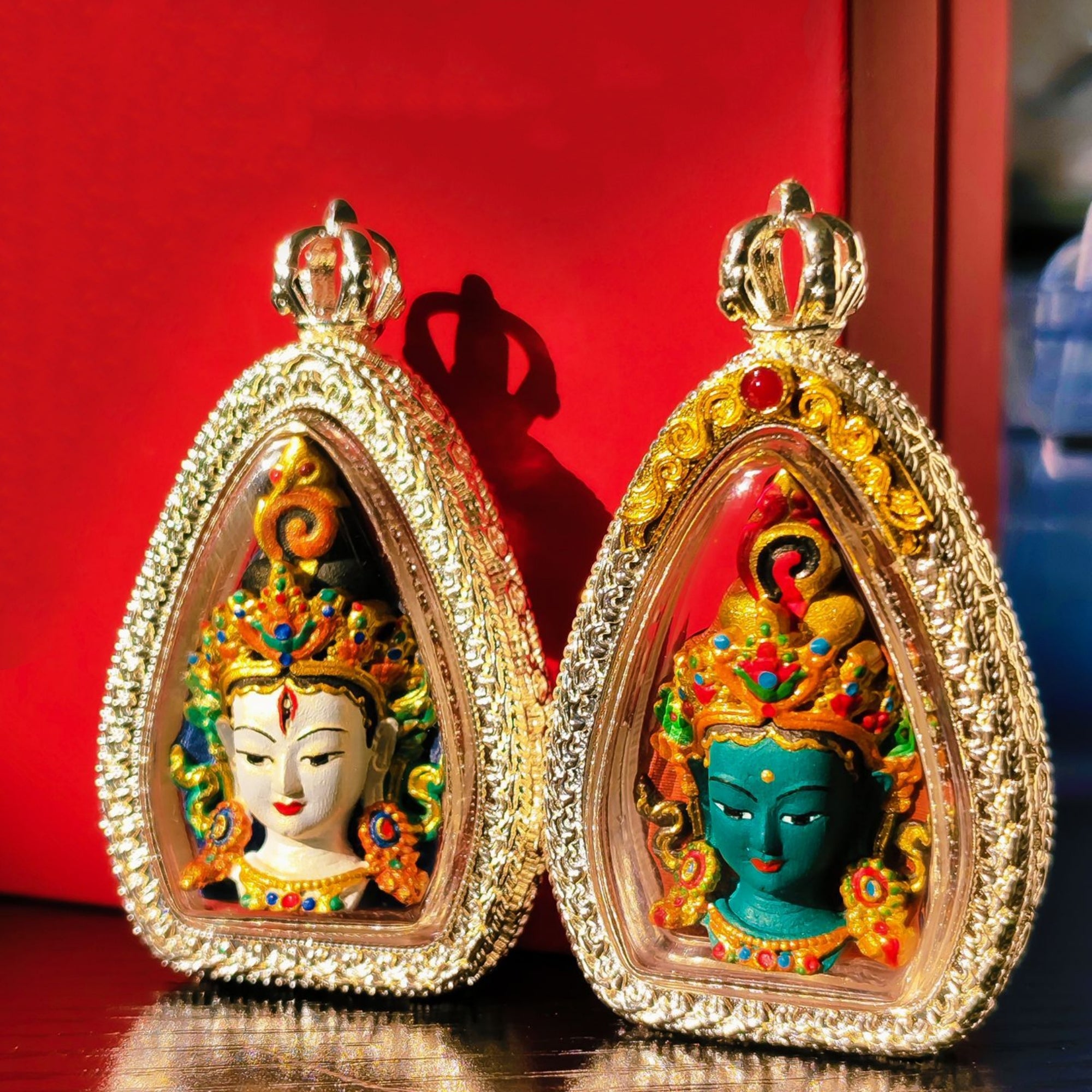 Two ornate teardrop-shaped pendant charms with crown tops, set against a red background. Each pendant features a colorful deity face under a clear dome, surrounded by a gold-toned beaded border.