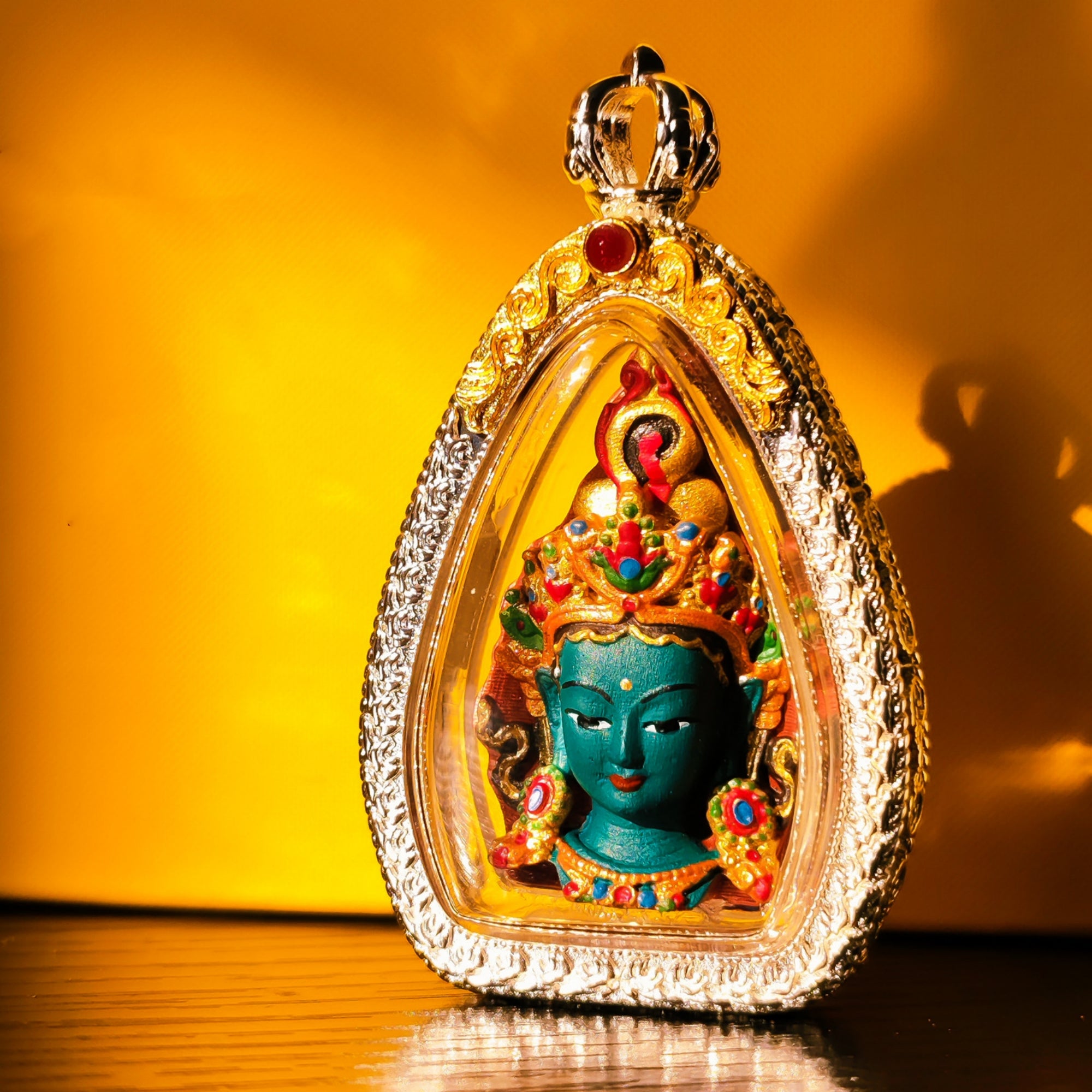 A teardrop-shaped ornate pendant or amulet featuring a turquoise-blue deity face surrounded by gold-toned detailing, red and green jewel accents, and a silver-toned decorative border, photographed against a warm yellow-orange background.