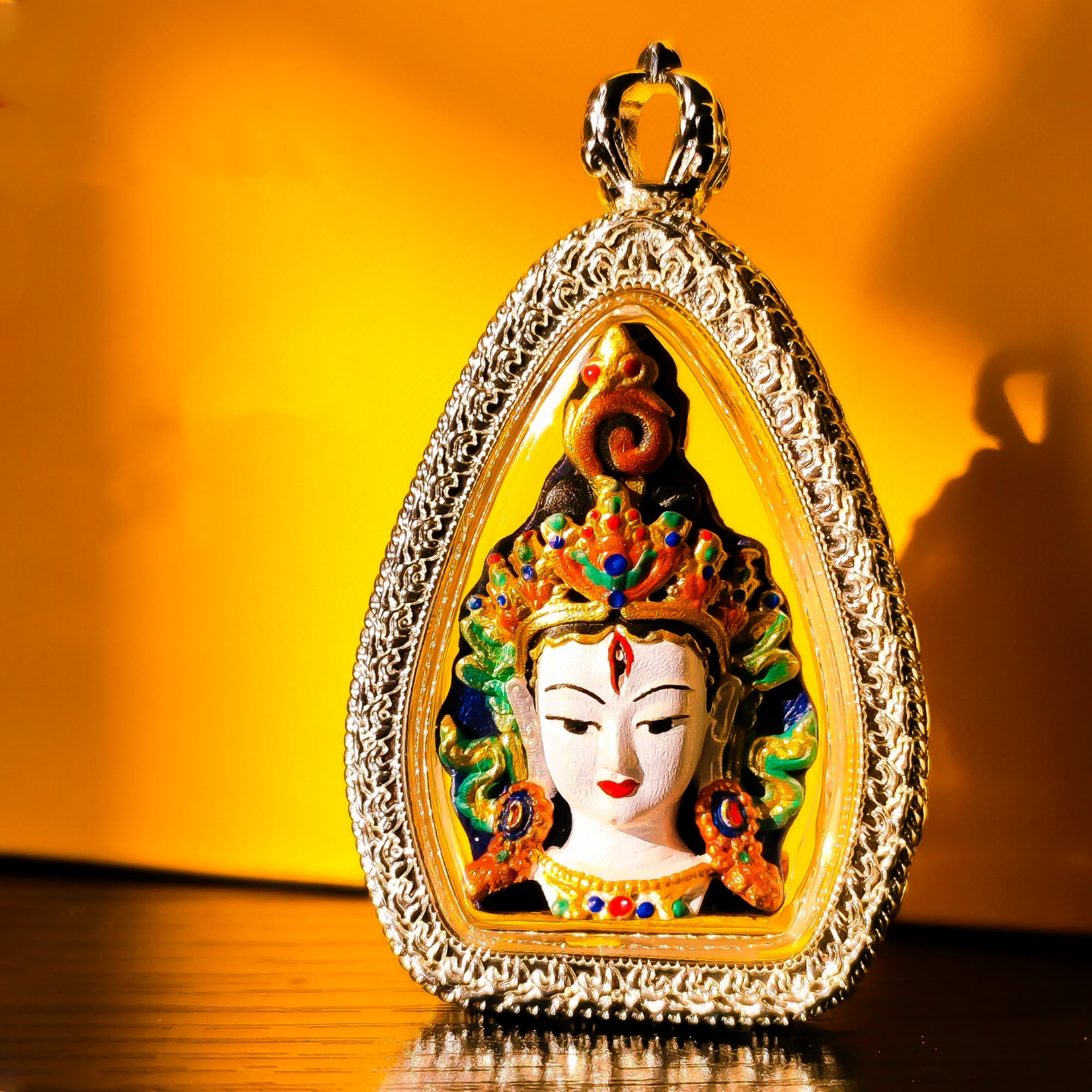 A teardrop-shaped ornate pendant featuring a colorful deity or Buddha face in the center, surrounded by a textured gold-toned frame and hanging loop, photographed against a warm glowing yellow-orange background.