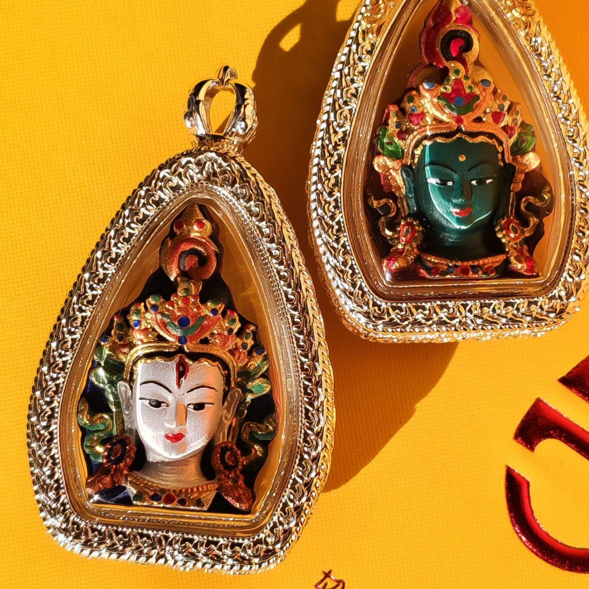 Two ornate teardrop-shaped religious pendant amulets with detailed enameled deity faces, displayed on a bright yellow background.