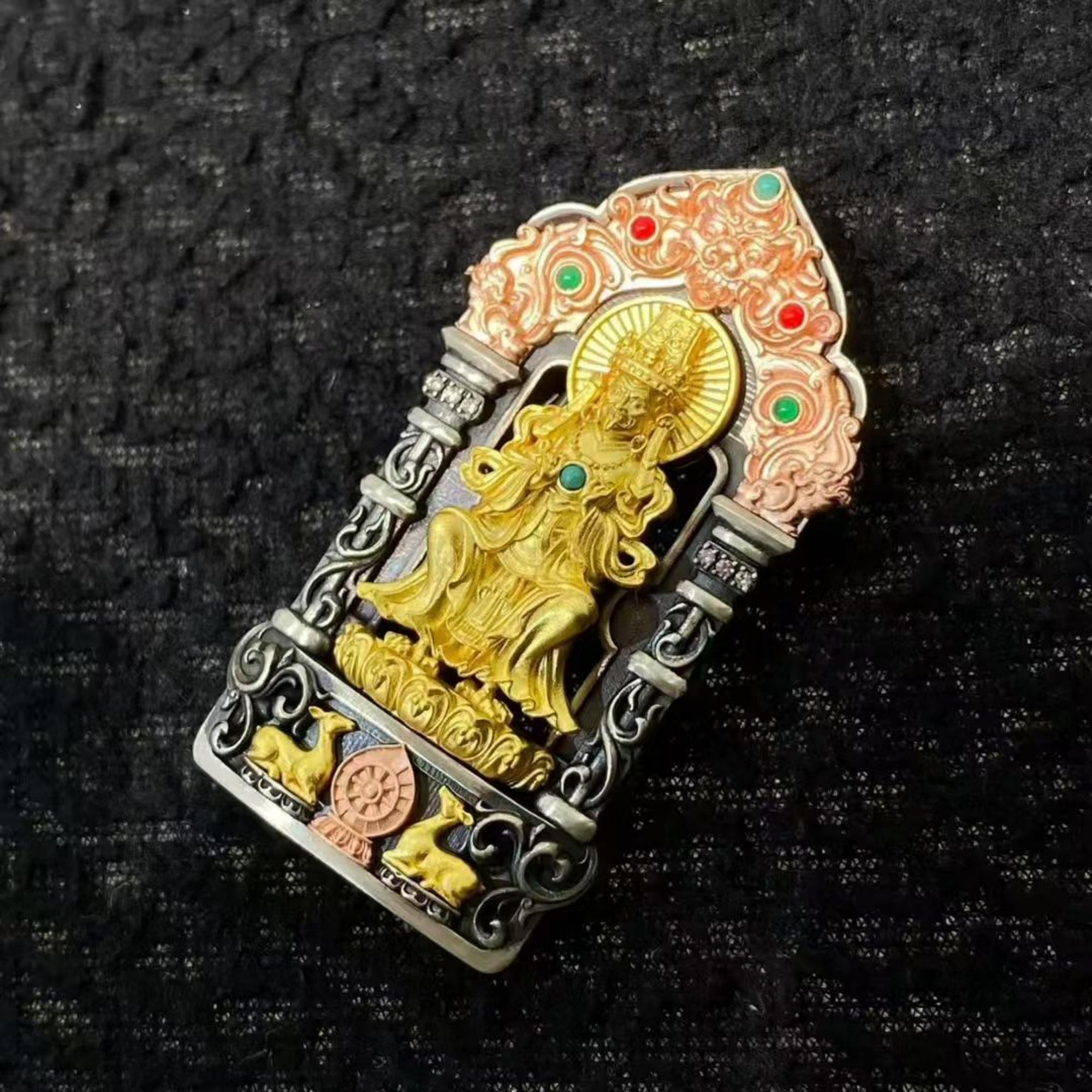 A decorative religious pendant or brooch featuring a gold-toned seated deity figure, surrounded by silver-toned ornate scrollwork and a peach-pink arched top with small red and green stone-like accents, photographed on a dark textured fabric background.