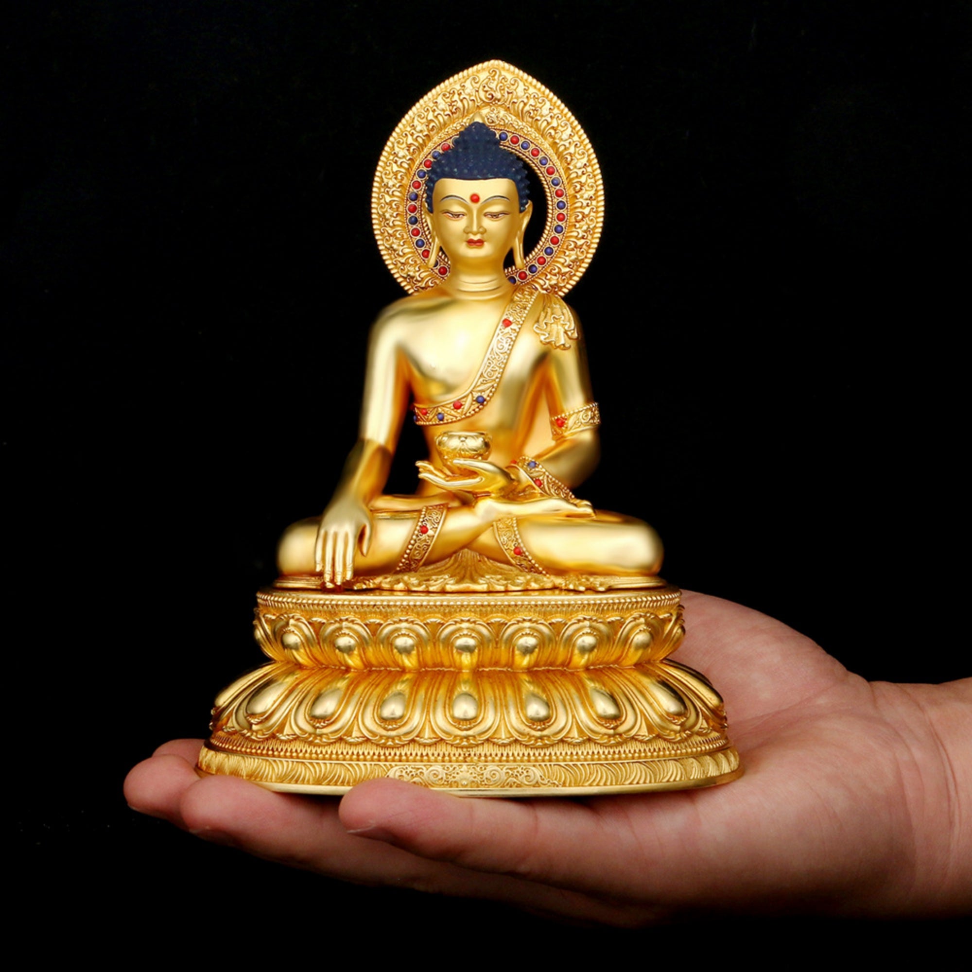 A small golden Buddha statue seated on a lotus base, held in a person's hand against a black background.