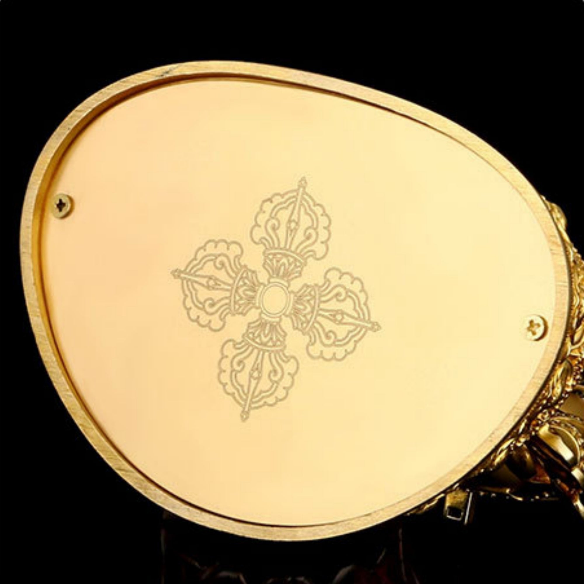 A close-up of a gold-toned oval plate or cover with a decorative engraved cross-like motif in the center, shown against a black background.