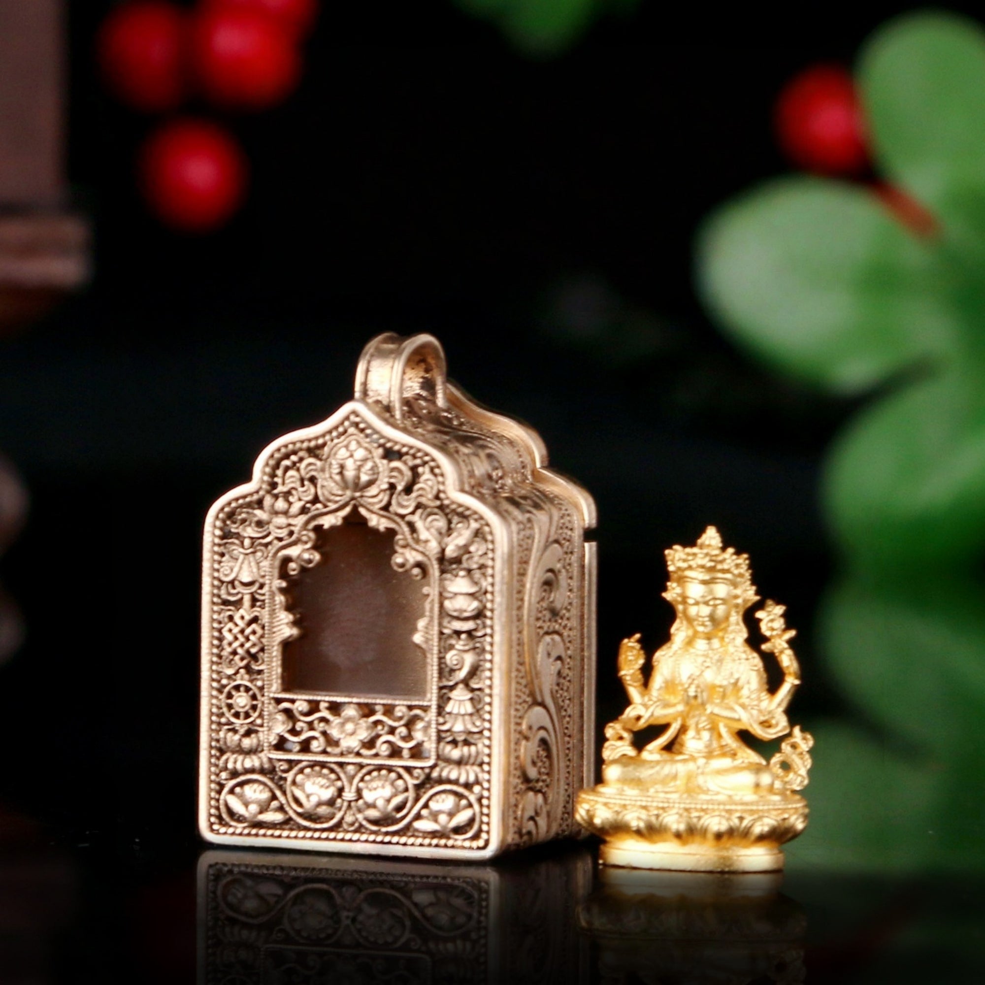 An ornate antique gold-toned metal temple box with intricate filigree detailing, shown next to a small golden deity figurine on a dark reflective surface with blurred green leaves and red berries in the background.