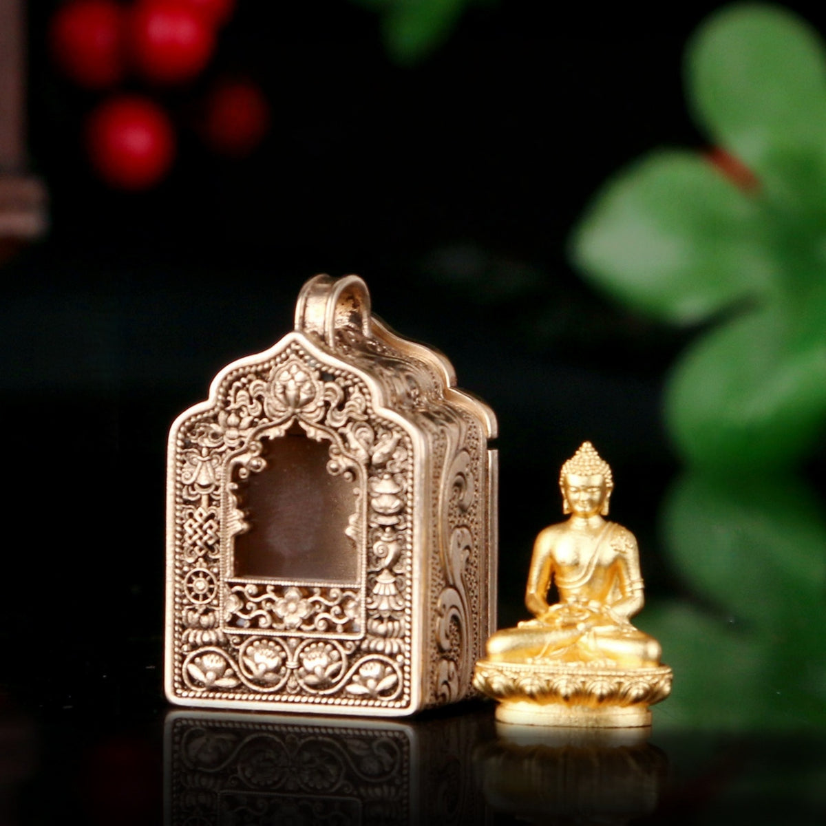 An ornate antique-style gold-toned metal box with intricate relief carvings and a small seated Buddha figurine beside it on a dark reflective surface, with blurred green leaves and red berries in the background.