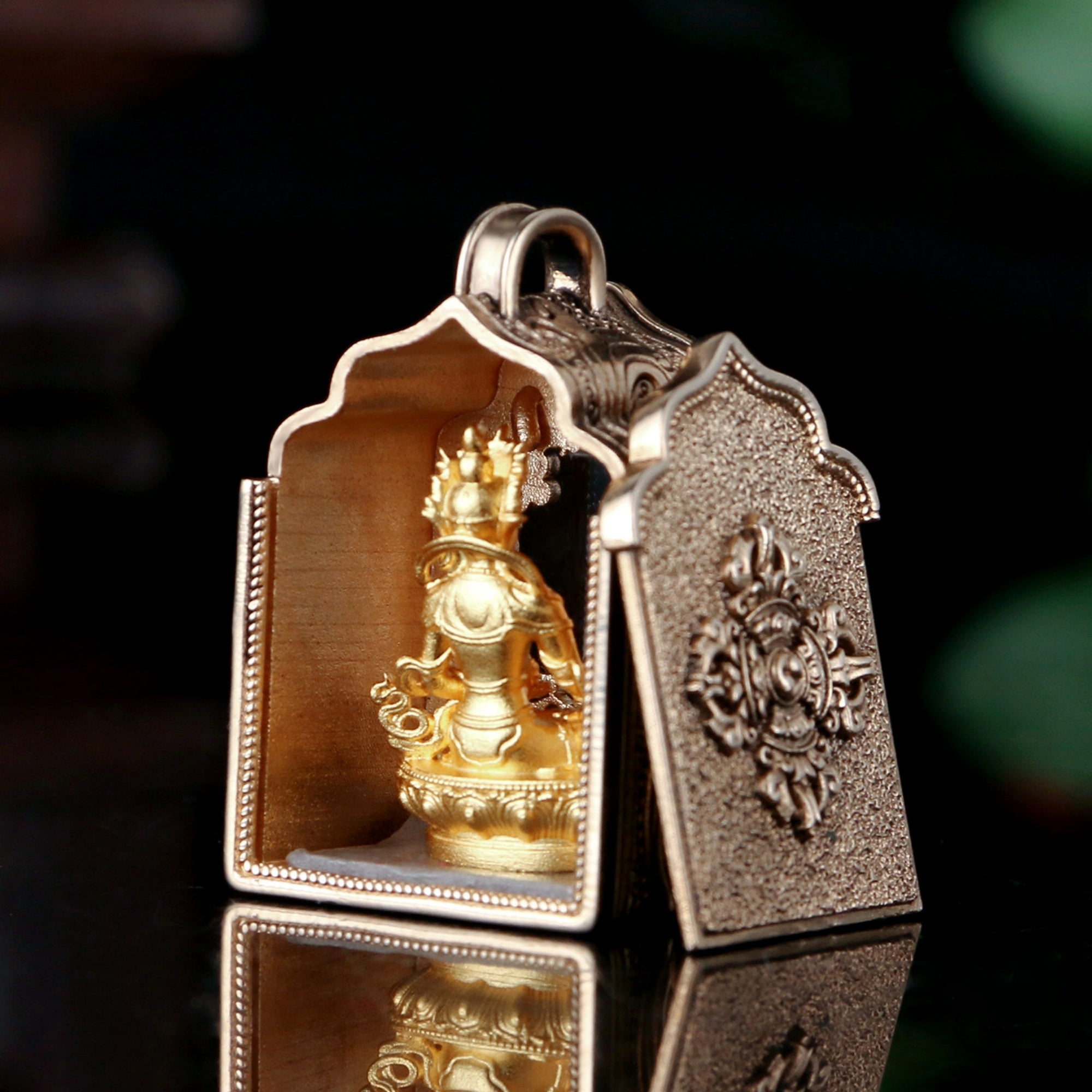 A small ornate metallic pendant or amulet with a hinged open-front design, featuring a gold-toned deity figure inside and a textured silver-toned outer panel with embossed decorative details, shown on a dark reflective surface.