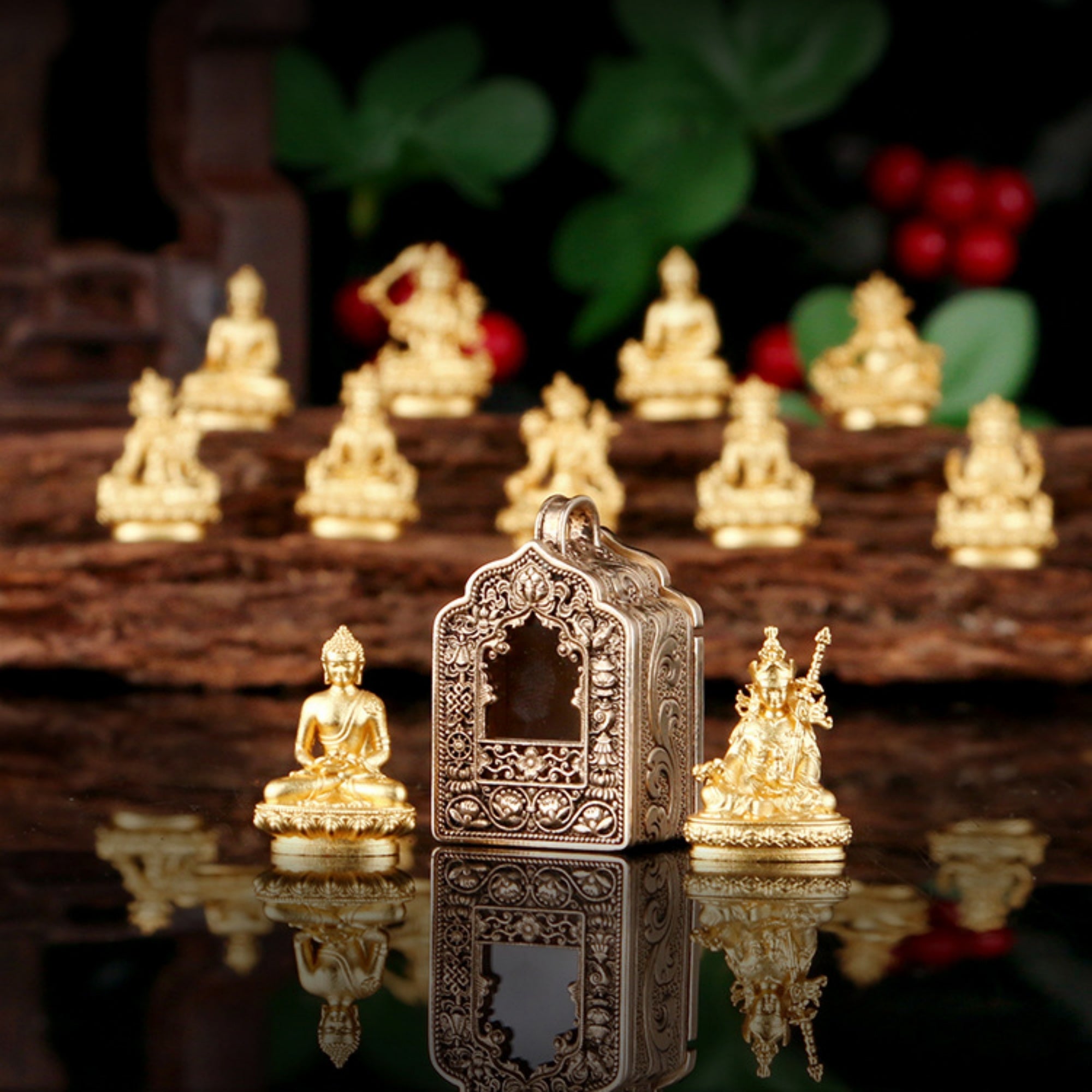 A decorative miniature Buddha and deity figurine set displayed on a reflective dark surface, with a central ornate silver-toned temple structure flanked by gold-toned seated figures. Smaller Buddha statues and red berries appear blurred in the background on a wooden surface.