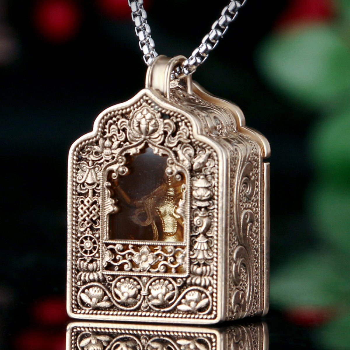 An ornate antique gold-toned pendant necklace featuring a detailed Ganesha design set in a box-like locket with a silver chain, shown against a dark, blurred festive background.