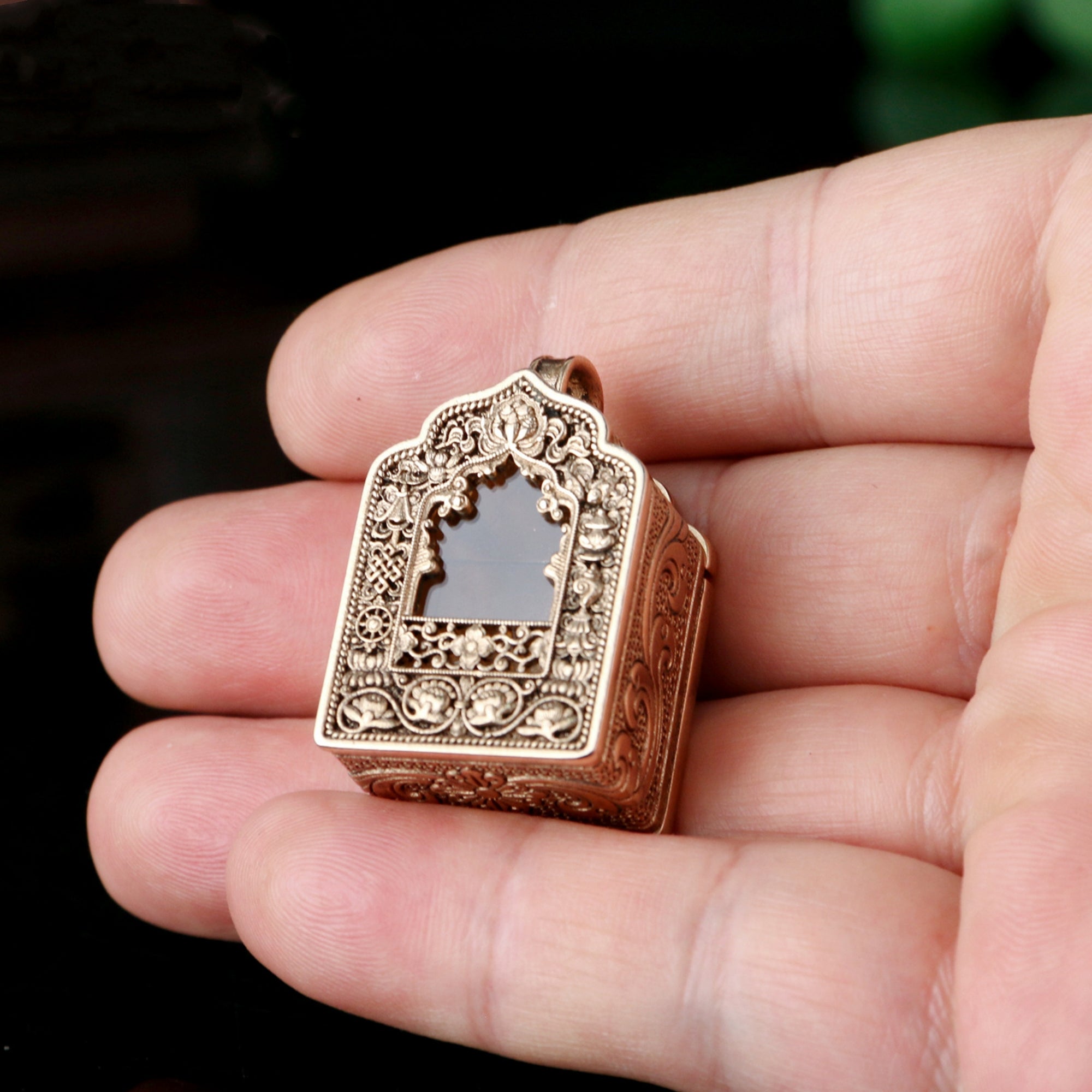 An ornate gold-toned metal pendant with intricate filigree detailing and a central dark inlay, held in a person's hand against a dark background.
