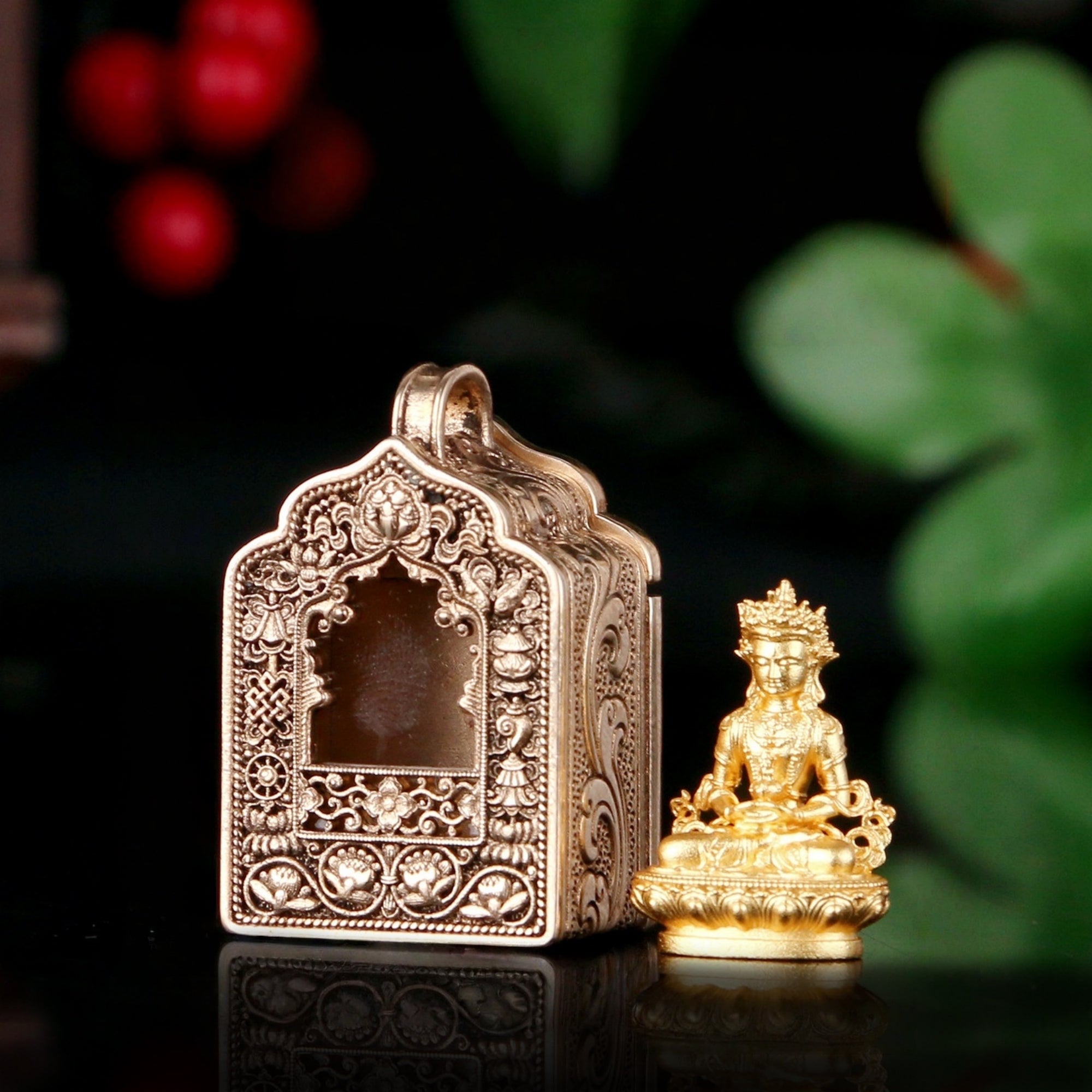 An ornate antique-style gold-toned metal box with intricate floral engravings and a small seated Buddha figurine beside it, set against a dark background with blurred green leaves and red berries.