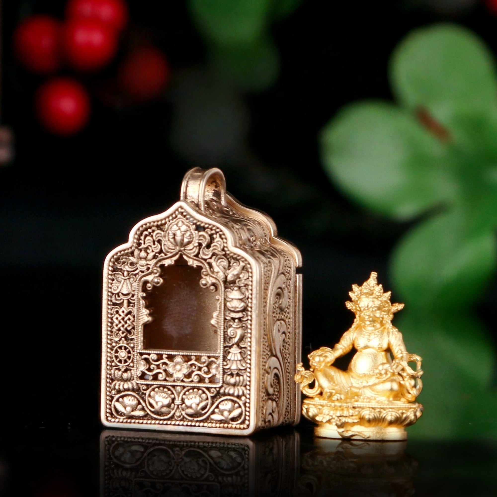 An ornate metallic box with intricate filigree detailing and a small seated deity figurine beside it, photographed on a dark reflective surface with blurred green leaves and red berries in the background.