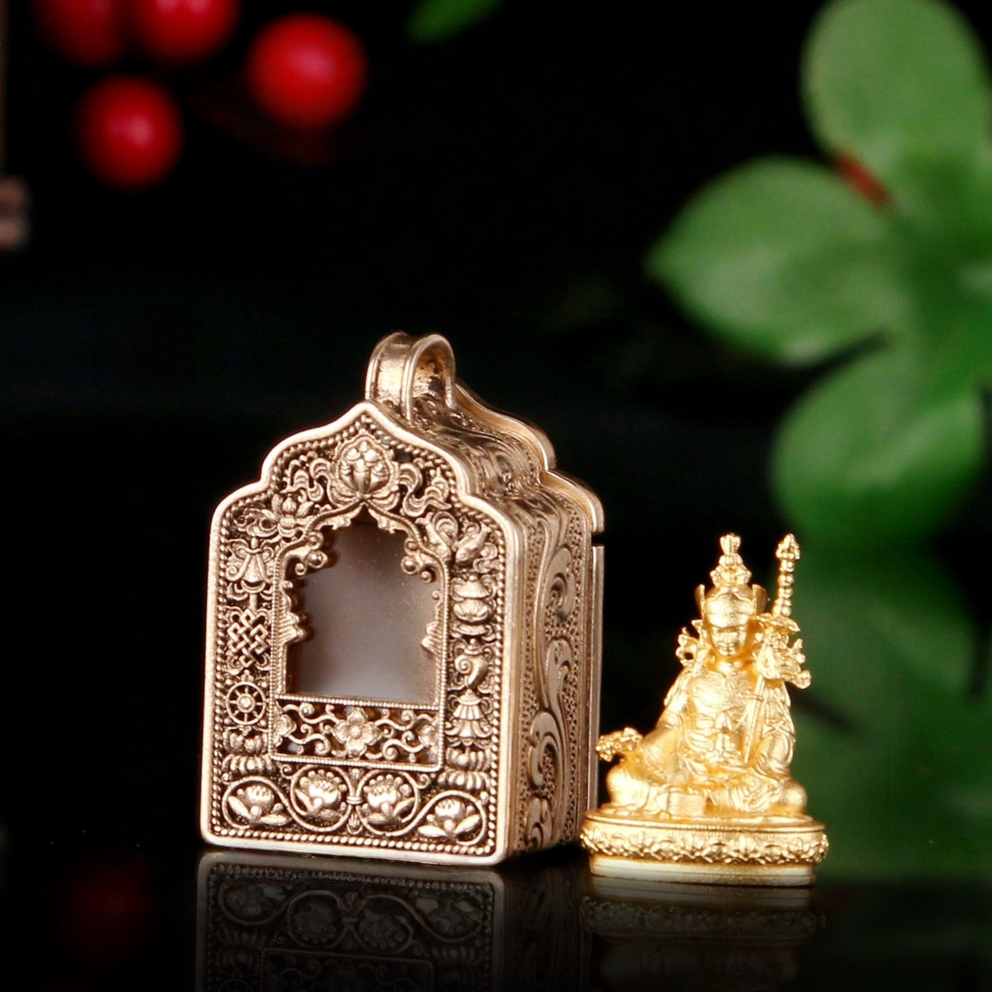 A small ornate gold-toned decorative box with intricate relief carvings and a hanging loop, shown beside a matching small seated figurine on a dark reflective surface with blurred red berries and green leaves in the background.