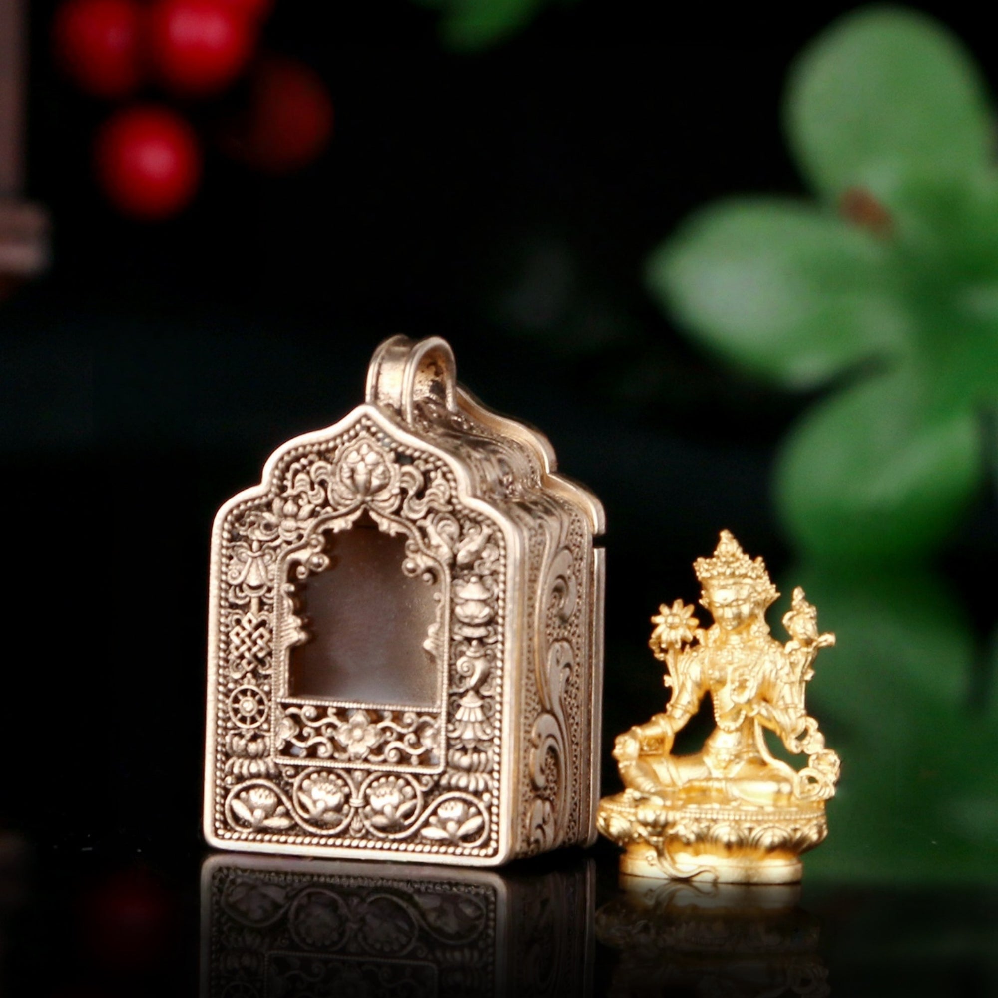 An ornate silver-toned metal box with intricate floral engravings and a small gold-toned deity figurine beside it, set against a dark background with blurred green leaves and red berries.