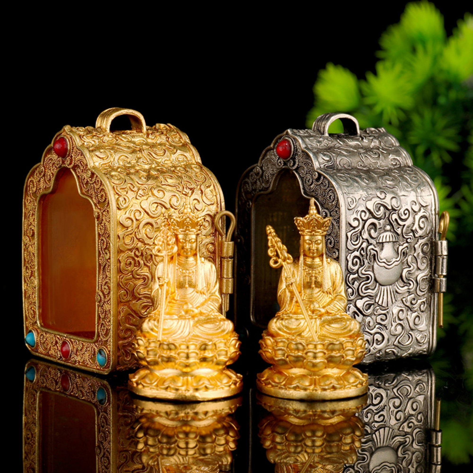 Two ornate decorative boxes with small seated deity figurines displayed in front, one in polished gold and one in silver-toned metal, set against a black background with green foliage at the upper right.