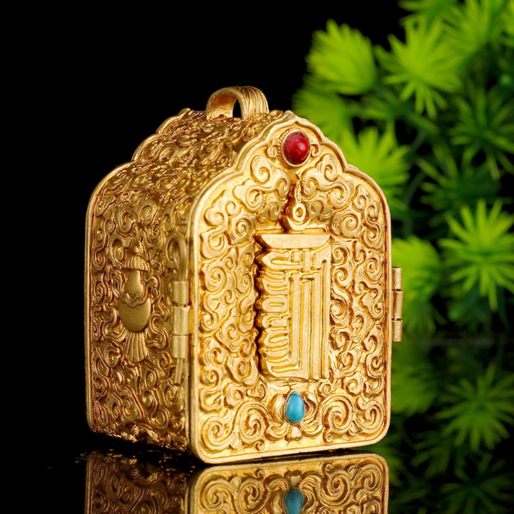 An ornate gold-toned decorative box with intricate embossed scrollwork, a central vertical relief, and small red and blue gemstone accents, photographed against a black background with blurred green foliage on the right.