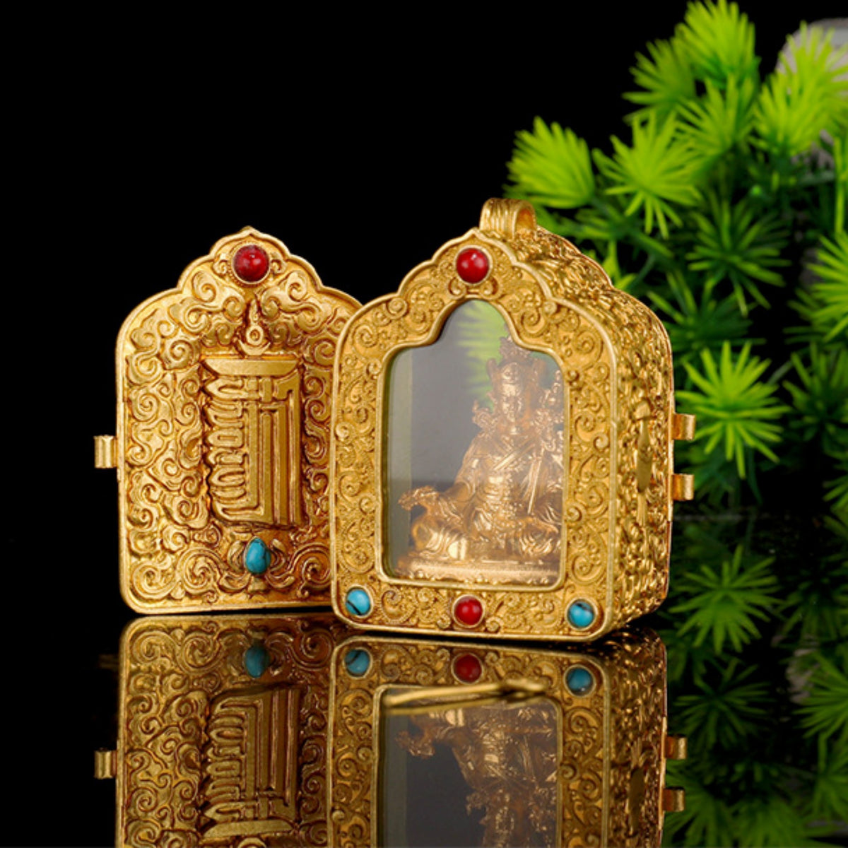An ornate gold-toned decorative box with carved detailing, a clear front panel showing a small figurine inside, and red and blue gemstone accents, photographed on a reflective black surface with green foliage in the background.