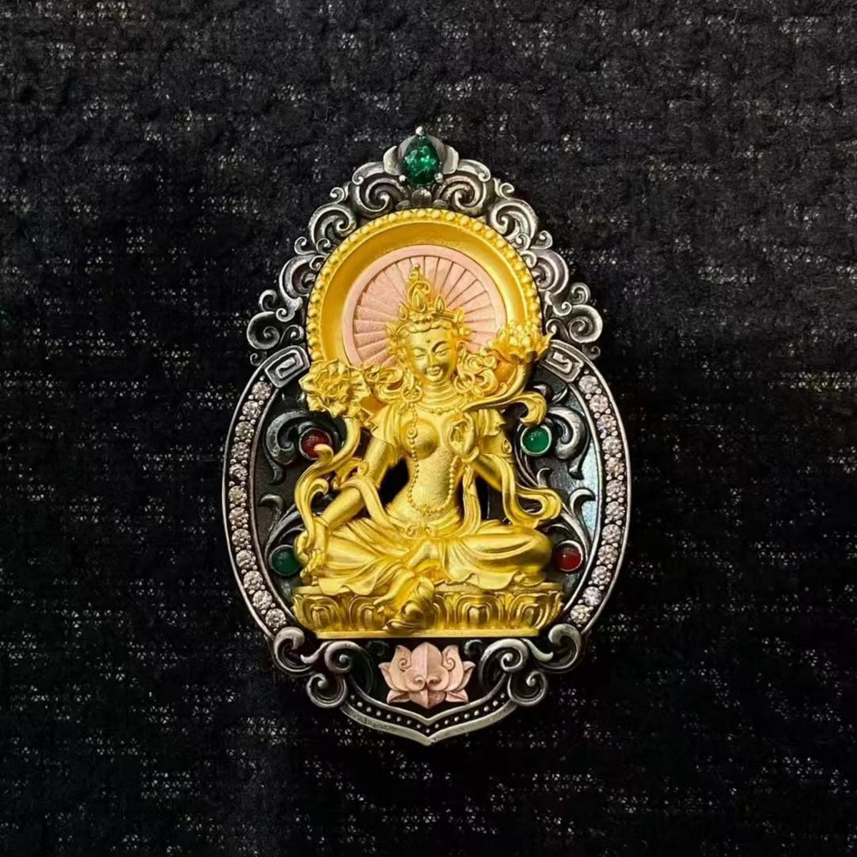 A detailed gold and silver oval pendant featuring a seated deity with green and pink gemstone accents, displayed on a textured black background.
