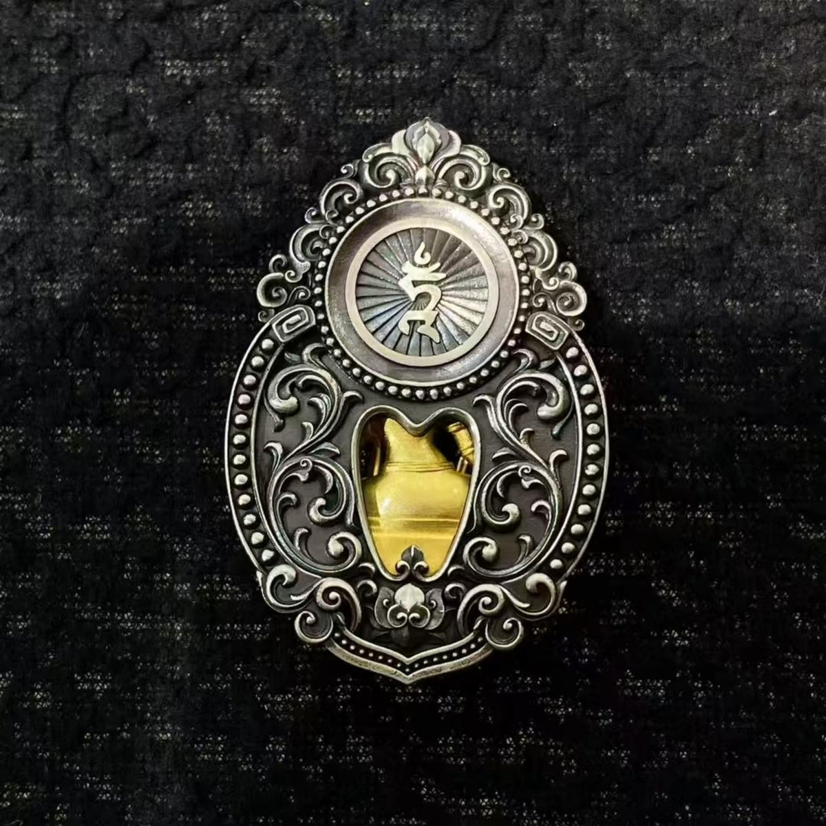 A silver-toned ornate pendant or brooch with a circular sunburst motif at the top and a central cutout revealing a yellow object inside, photographed against a dark textured fabric background.