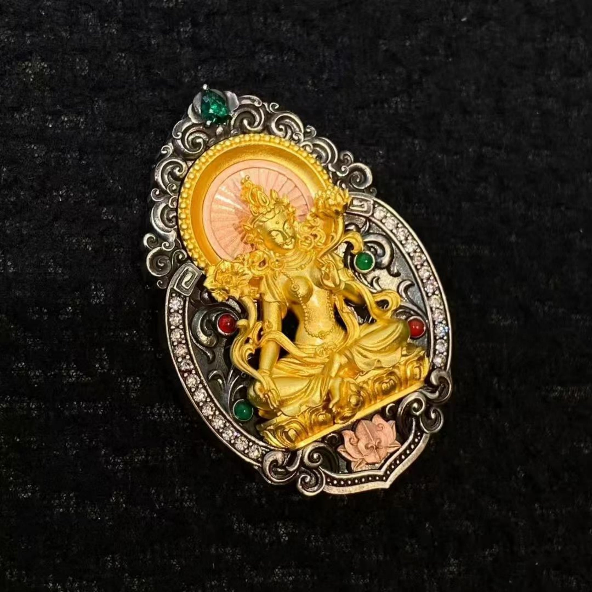 An ornate oval brooch featuring a detailed gold-toned seated deity figure, surrounded by silver-toned scrollwork and accented with small green and red gemstones on a textured black background.
