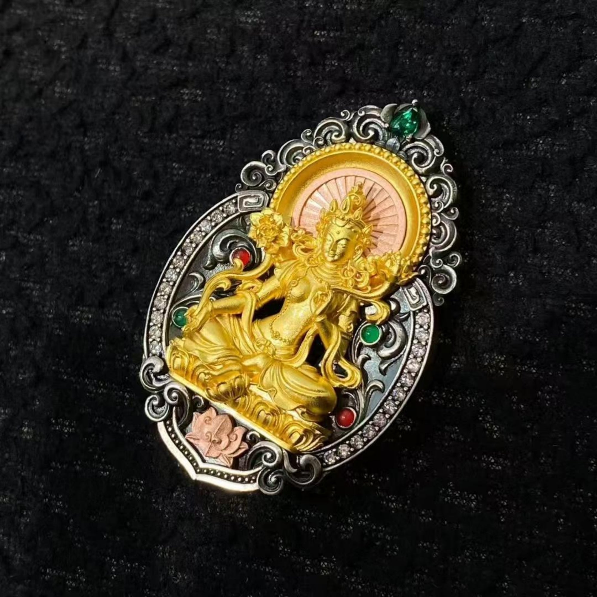 A gold and silver oval pendant featuring a seated multi-armed deity figure, accented with small green and red gemstones on a textured black background.