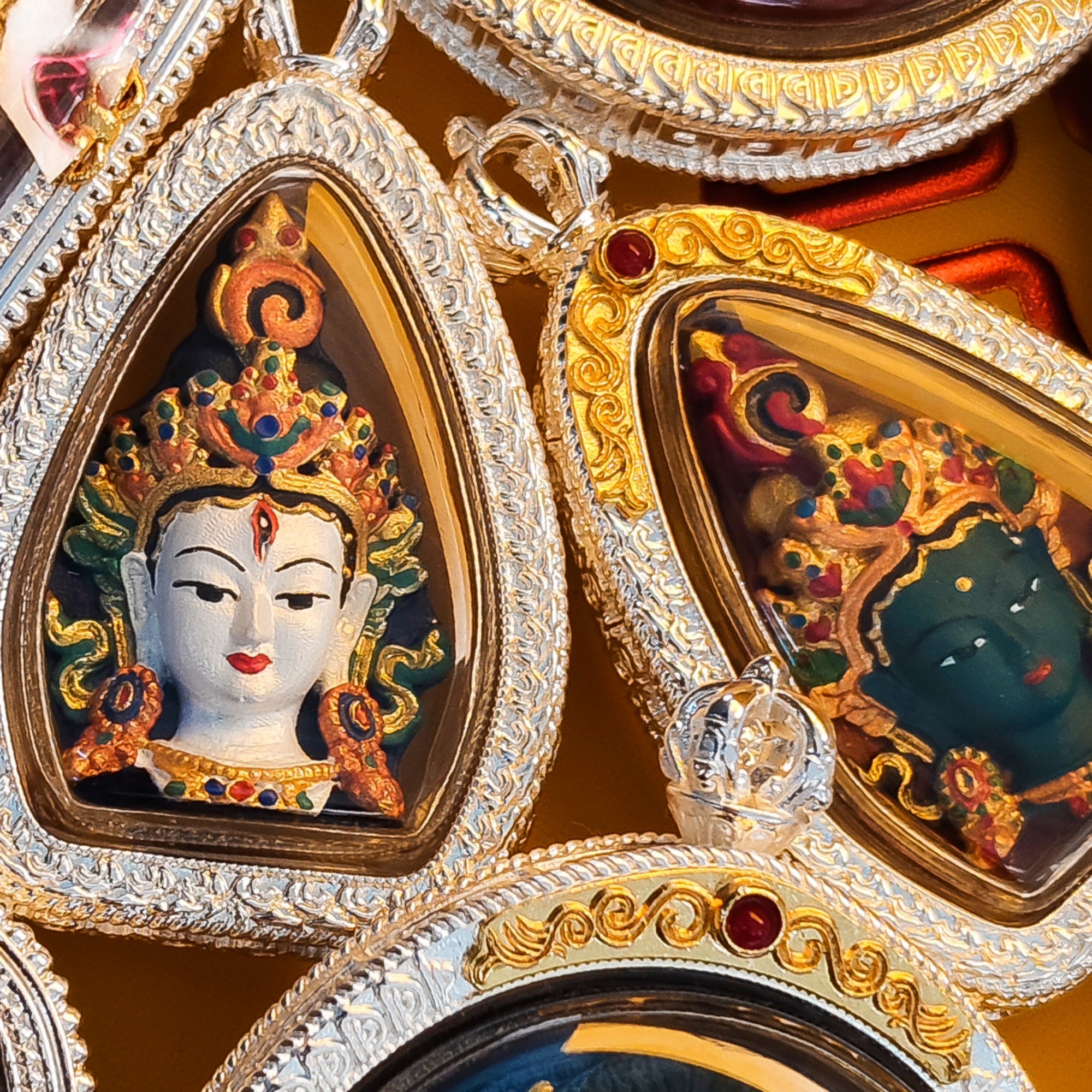Close-up of ornate teardrop-shaped religious amulets with detailed deity carvings under clear domes, set in silver and gold-toned engraved frames with small red gem accents.