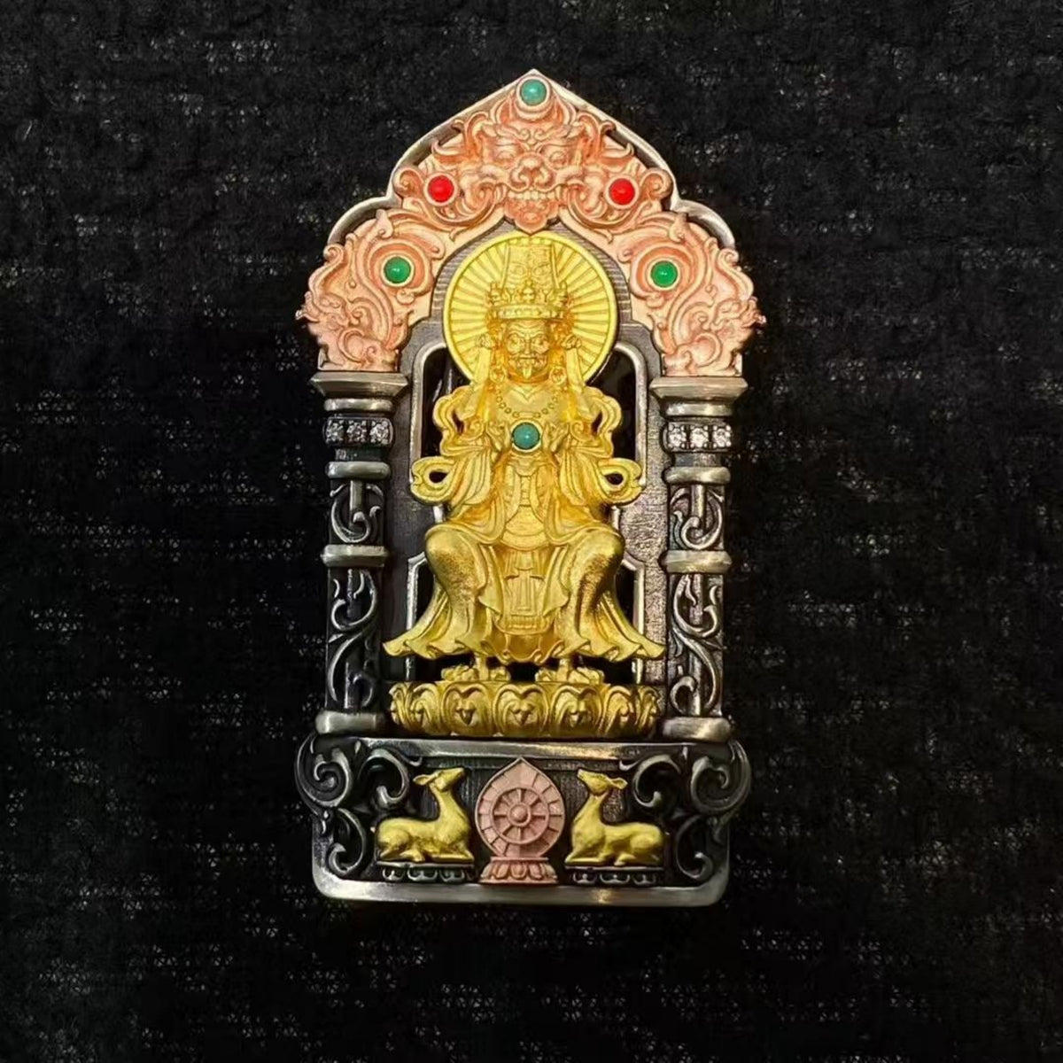 A decorative religious figurine or pendant featuring a seated golden deity in a silver-toned ornate frame with small red and green stone accents, photographed against a dark textured background.
