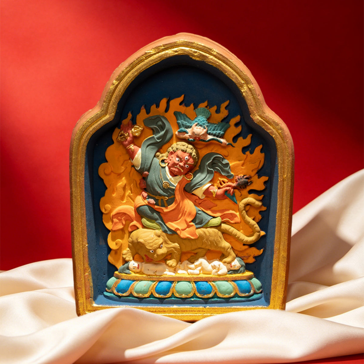A small arched decorative figurine or plaque with a gold-toned frame, featuring a colorful relief scene of a figure on a tiger against blue and orange flames, displayed on draped cream fabric with a red background.