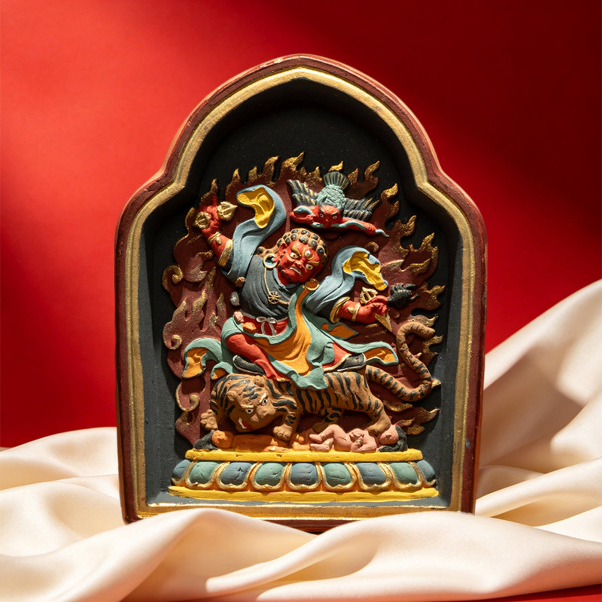 A small arched religious figurine depicting a warrior riding a tiger, shown on draped cream fabric against a red background.
